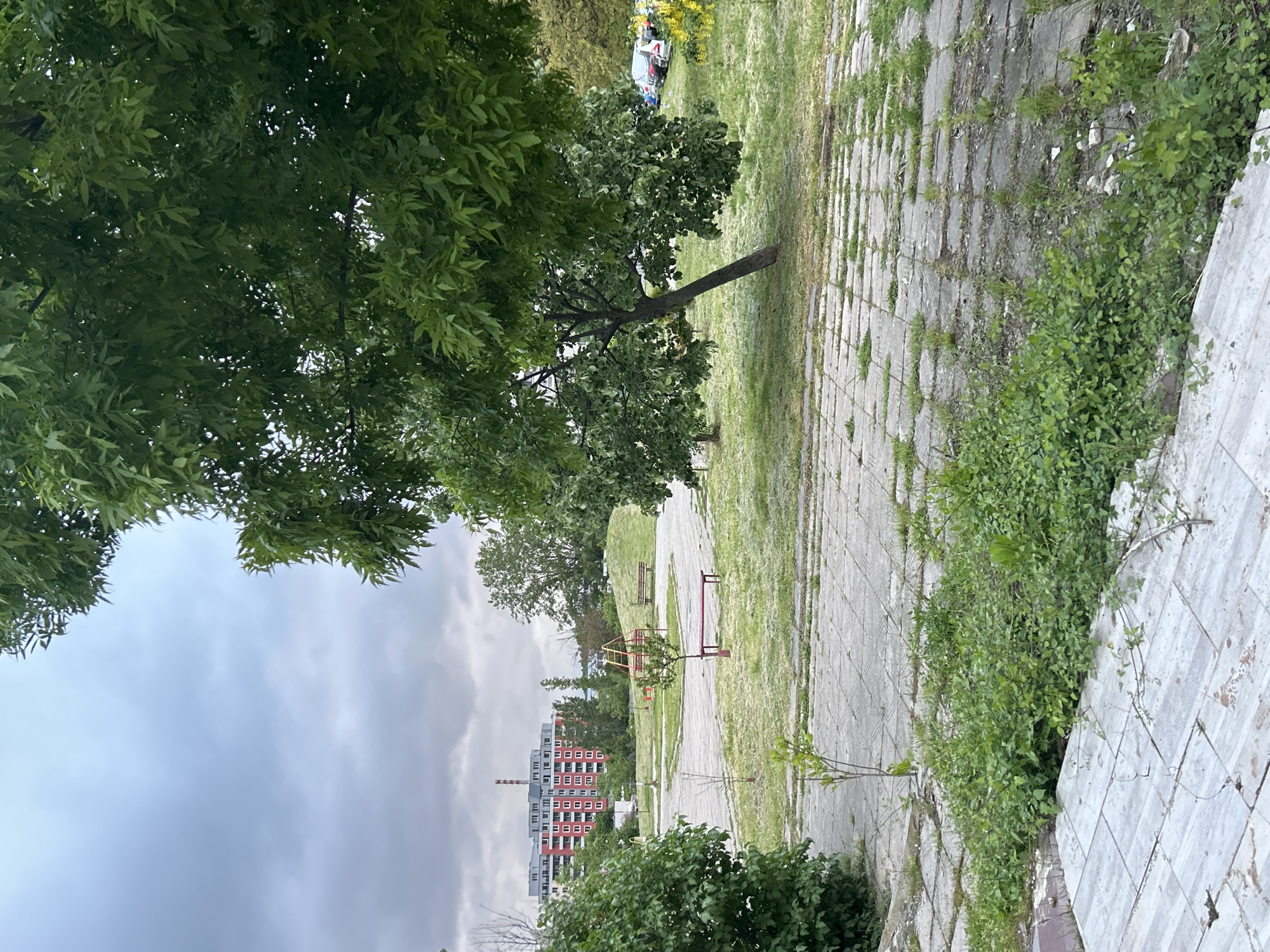 The image depicts a city park in an unspecified location. A paved path made of square stones leads into the distance, flanked by grass and greenery. A small tree grows at an angle from the left side of the path. Further along the path, a red metal structure resembling a playground element is visible. In the background, an apartment building with a chimney rises against a cloudy sky. The scene suggests an urban environment with a park area integrated into the cityscape.