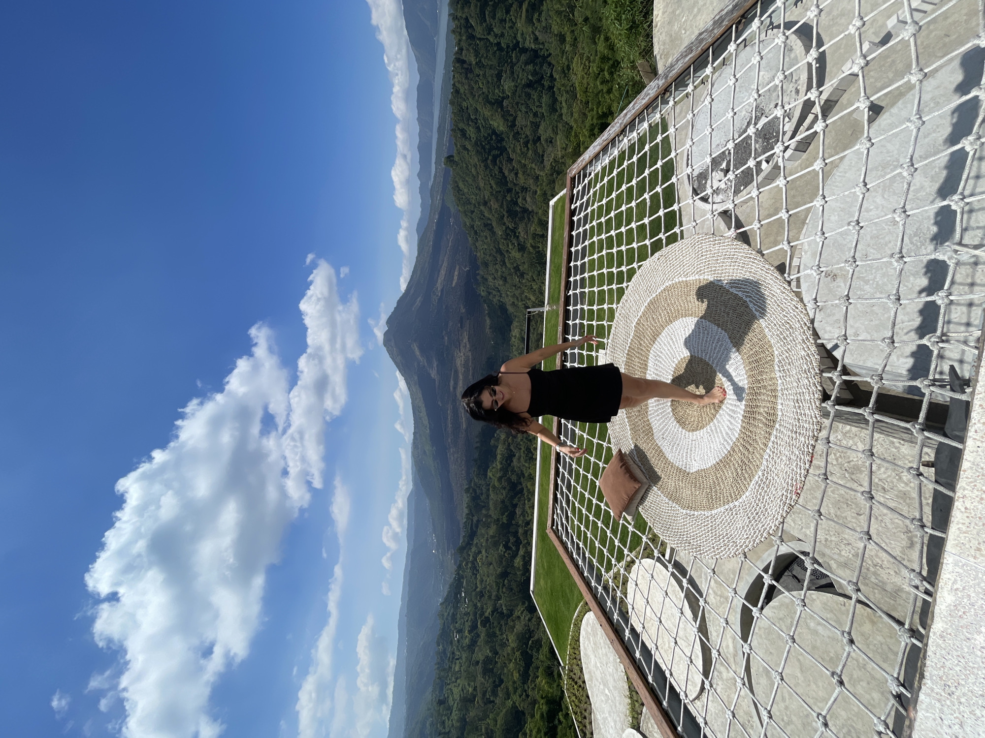 A woman in a black slip dress and sunglasses stands on a netted platform in Kintamani, Indonesia, on a clear, sunny day with fluffy clouds in a bright blue sky. The platform, made of white rope, has a circular woven mat in earthy tones and a brown cushion. Behind the platform, lush green trees cover rolling hills, leading to a volcanic mountain in the distance with a body of water beside it. The woman is looking down and to her right, with her arms slightly out. The scene suggests a tourist viewpoint with a panoramic natural landscape.