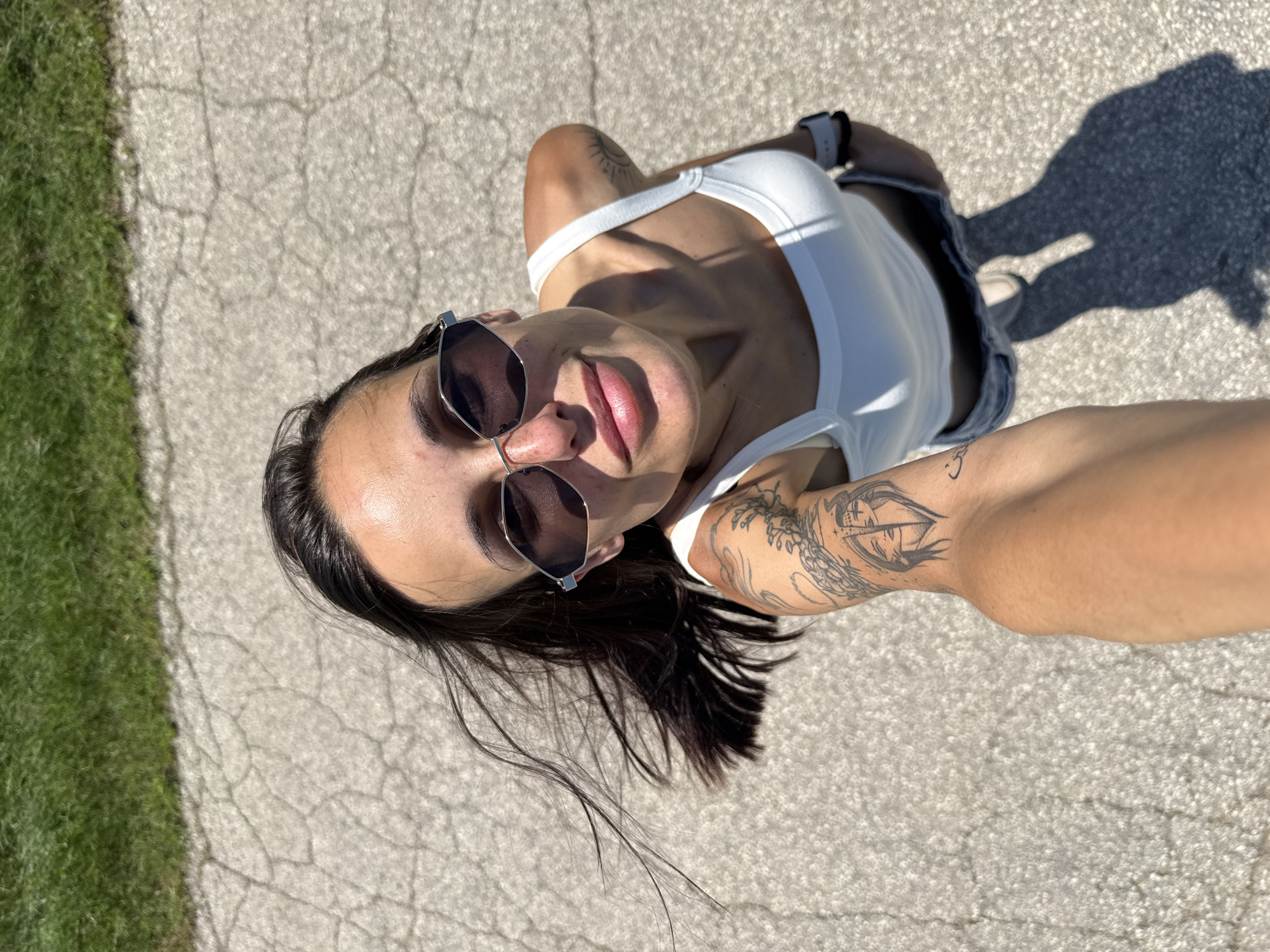 A high-angle selfie captures a young adult female on a cracked asphalt surface in Slavovitsa, Bulgaria. The subject, positioned centrally, has dark hair, partially windblown, and wears hexagonal-framed dark sunglasses. She exhibits a subtle smile with visible lips. Her attire consists of a white tank top and light-colored bottoms. Tattoos are visible on both arms: a detailed design featuring a stylized face split vertically on her left bicep, intricate floral patterns on her left upper arm, a small crescent-like symbol on her left forearm, and a minimalist sun outline on her right shoulder. A white-banded watch is present on her right wrist. Strong sunlight from the upper right casts distinct shadows, including the elongated shadow of her lower body and legs on the pavement. In the background, beyond the cracked pavement, is a strip of vibrant green grass.