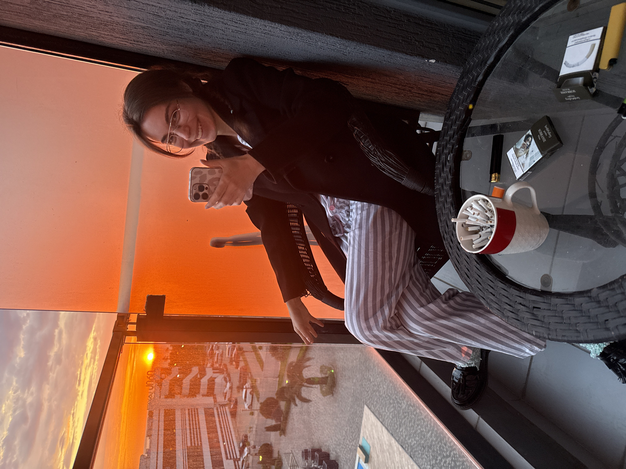 A woman wearing glasses, a black coat, and striped pajamas sits on a chair on a balcony. She holds a phone in her hand and smiles towards the camera. The balcony offers a view of the city of Batumi, Georgia, at sunset. On a table next to her are cigarettes in a cup, a lighter, and cigarette packs. The scene suggests a relaxed and casual atmosphere.