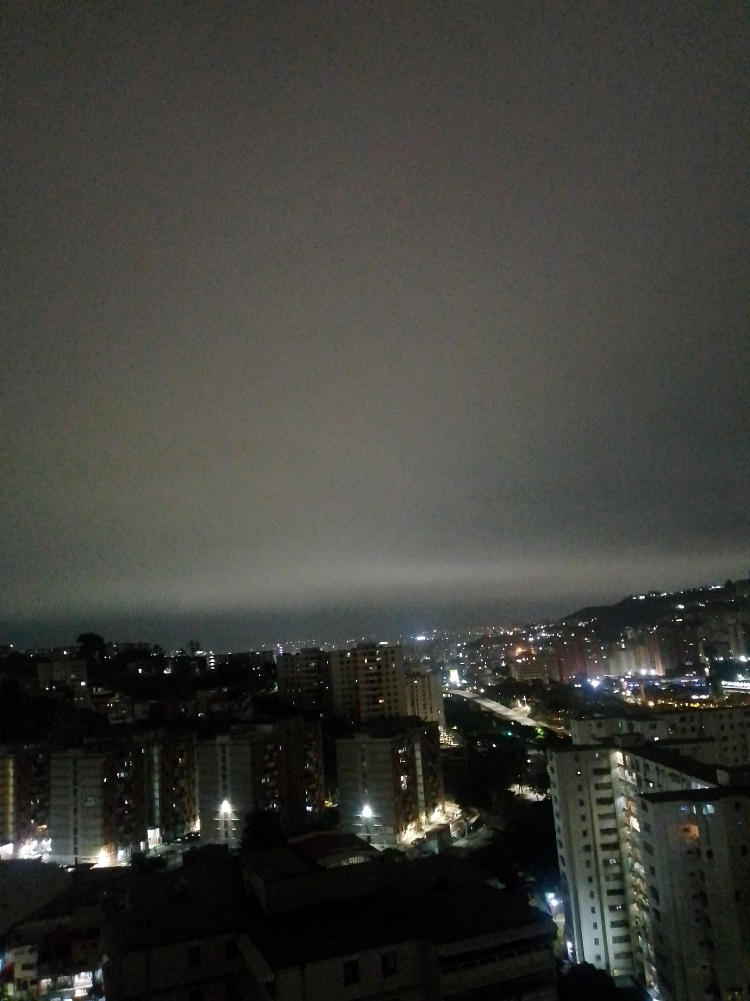 The image shows an aerial view of Caracas, Venezuela at night. Dense residential buildings with illuminated windows dominate the foreground and midground. A highway with lights can be seen winding through the city. In the distance, hills are dotted with lights. A heavy, overcast sky obscures the upper portion of the image.