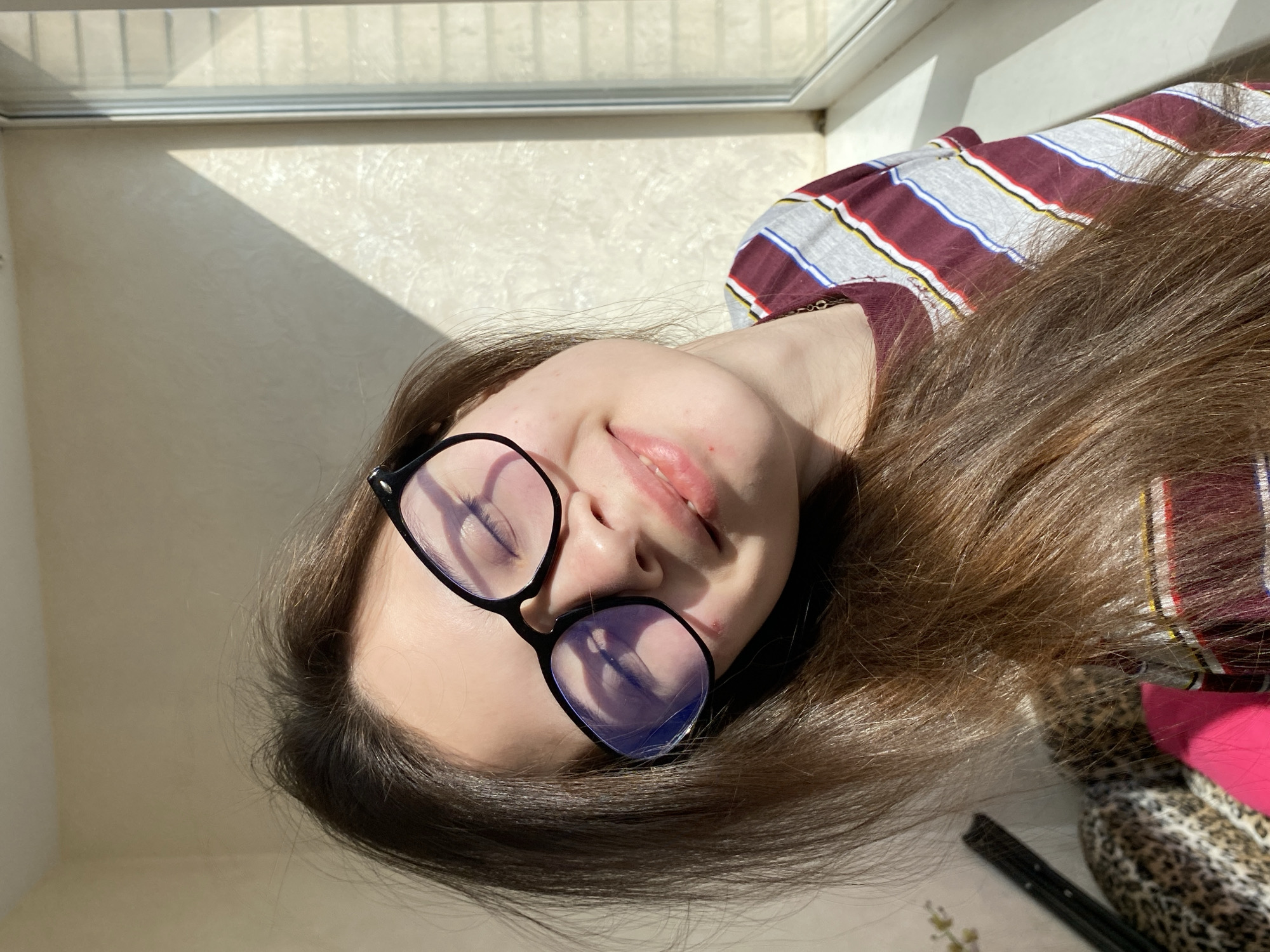 A candid, sunlit photograph features a young person, possibly female, lying down and looking upwards. Their head is positioned on the left side of the frame, with long, dark brown hair fanning out to the right. They are wearing black-rimmed glasses, and their eyes are closed or mostly closed, conveying a sense of peaceful relaxation. A gentle smile plays on their lips.
The strong, natural sunlight streams in from a window, casting crisp, angular shadows onto the light-colored, textured wall, suggesting a bright and sunny daytime setting. The window frame itself is visible along the top edge of the image. The person is dressed in a striped shirt with maroon, white, blue, and yellow lines. Partial views of a leopard print fabric and a bright pink item are visible near their hair on the right, along with a dark, slender object, possibly a tool or accessory. The reflections of the bright window are noticeable on the lenses of their glasses. The indoor scene, with its strong natural light, is set in Votkinsk, Russia. There is no visible text in the image.