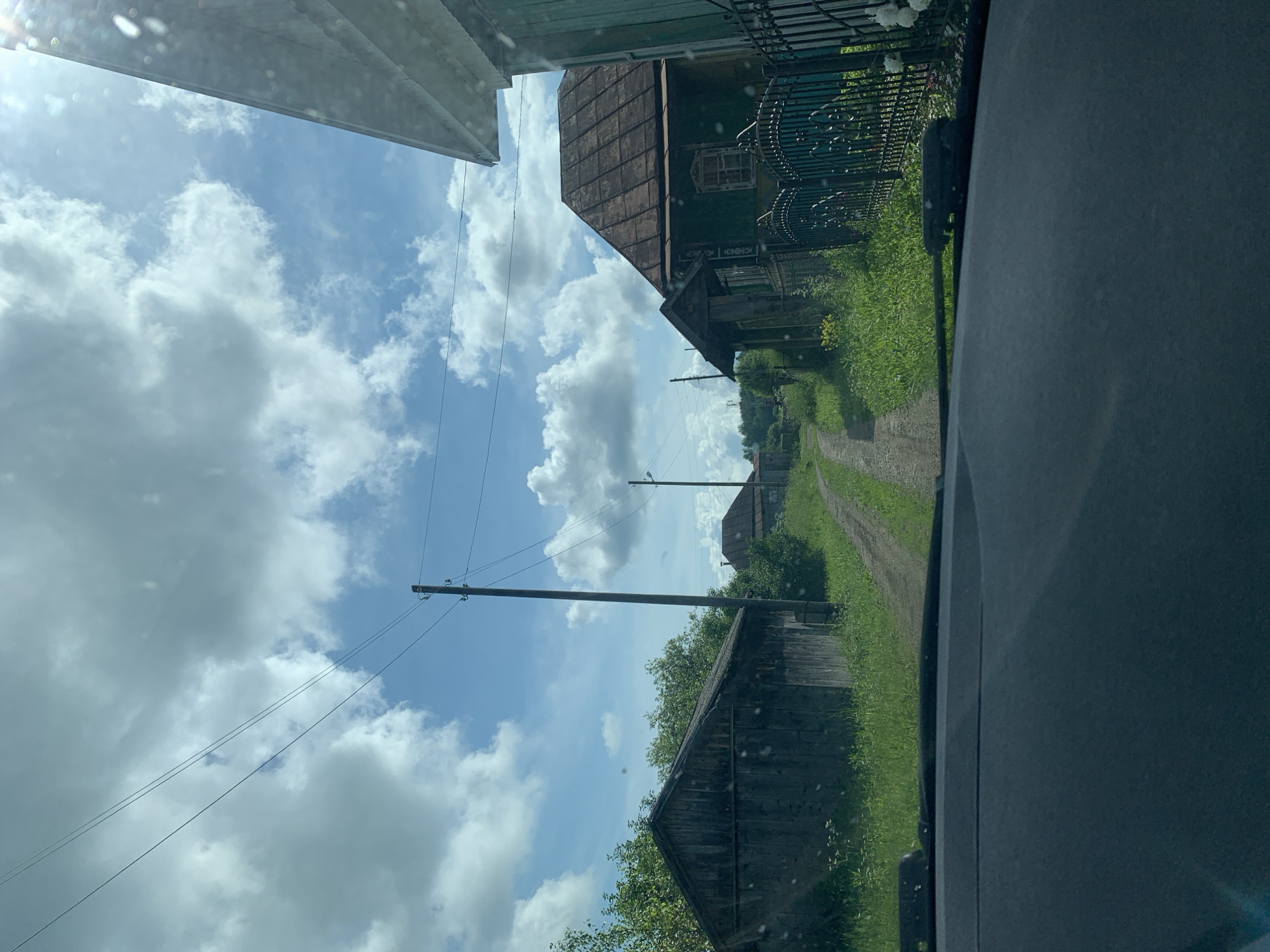 The image shows a rural scene in Barsai, Russia. The perspective is from inside a vehicle, looking out through the windshield. The sky is a vibrant blue with scattered white, fluffy clouds, suggesting a sunny day, likely mid-morning or afternoon.
The foreground is dominated by the dark interior of the vehicle, with a textured dashboard on the right. A dirt road, lined with lush green grass and foliage, curves into the distance. Along the road, several wooden structures are visible. On the left side of the road, a tall wooden utility pole with wires stretches across the frame. Further down the road, a traditional-looking wooden house with a dark, gabled roof and a decorative window frame can be seen. Behind this house, a dark metal fence with an ornate design runs along the property. Another wooden outbuilding, possibly a shed or barn, with a weathered appearance, is located closer to the foreground on the left.
The overall impression is one of a peaceful, rural setting, possibly a village or a countryside road. The scene conveys a sense of calm and natural beauty. There is no visible text in the image.