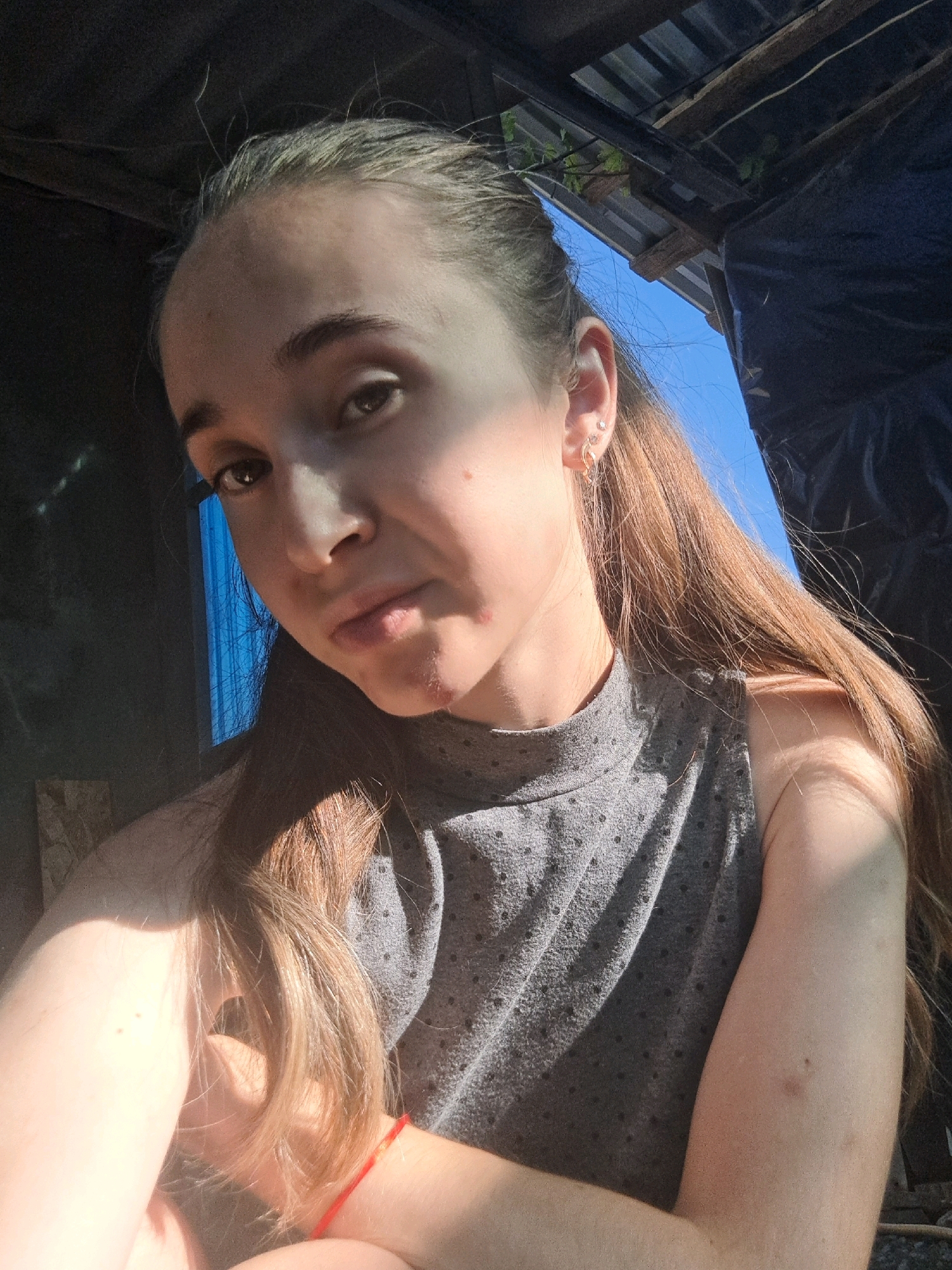 The image shows a young woman with long, light brown hair pulled back into a ponytail, wearing a grey sleeveless top. She is looking at the camera with a neutral expression. Her left arm is bent across her chest, and her right arm is visible on the right side of the frame. The background appears to be outdoors, possibly in a rural or semi-rural setting, with a blue sky visible above and some corrugated metal roofing. The lighting suggests it is daytime, with strong sunlight casting shadows. The woman has a small pimple on her chin. The location is Vypasne, Ukraine.