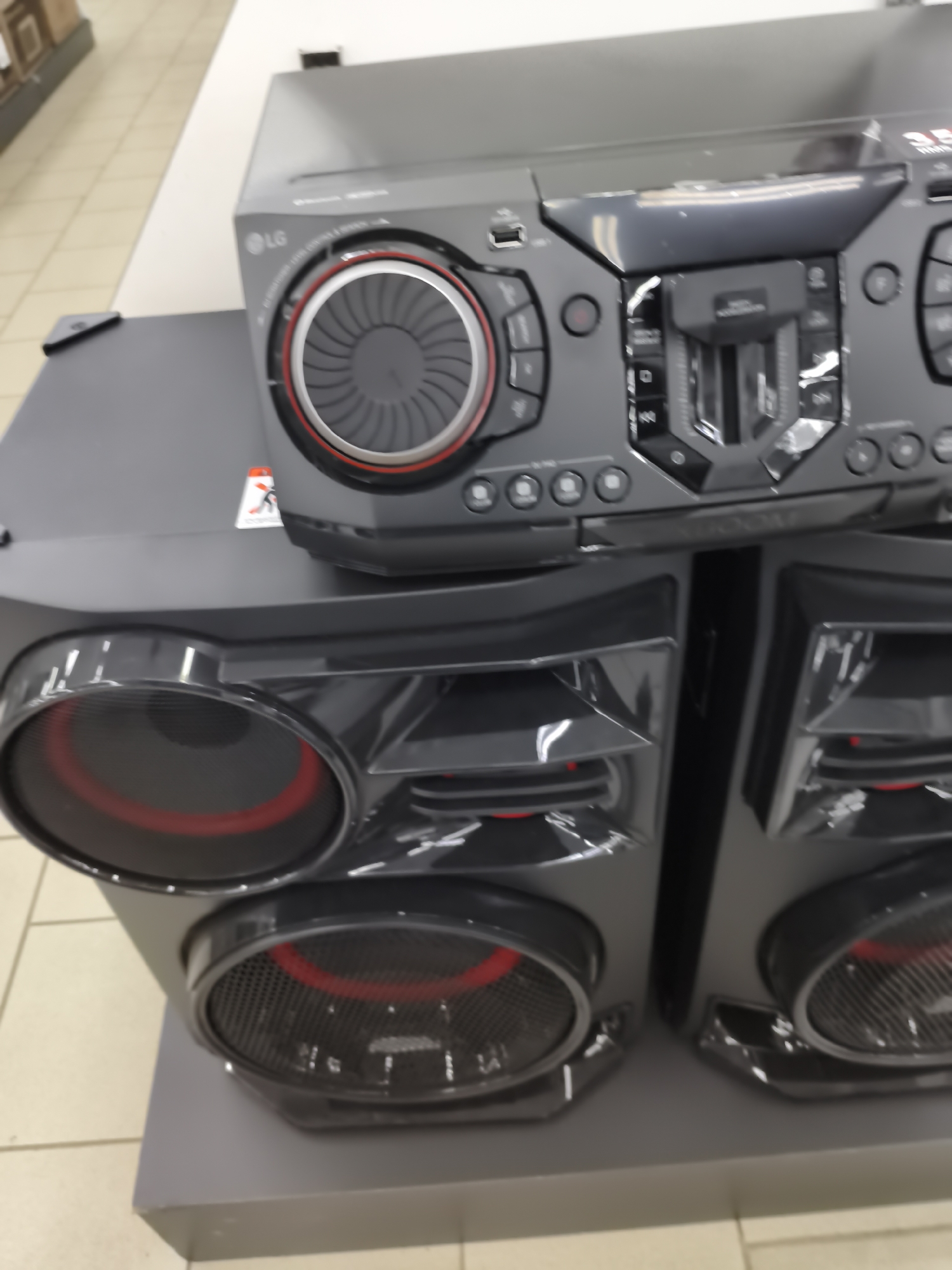 The image shows a close-up view of a black LG sound system, likely in a retail store setting in Aktobe, Kazakhstan. The sound system features two large speakers and a central unit with a CD player, control buttons, and a USB port. The speakers have black grilles with red accents around the circular openings. The central unit has a glossy black finish with various buttons and a recessed CD tray. The background shows a tiled floor and what appears to be part of a wall or display shelf. There are no people visible in the image.