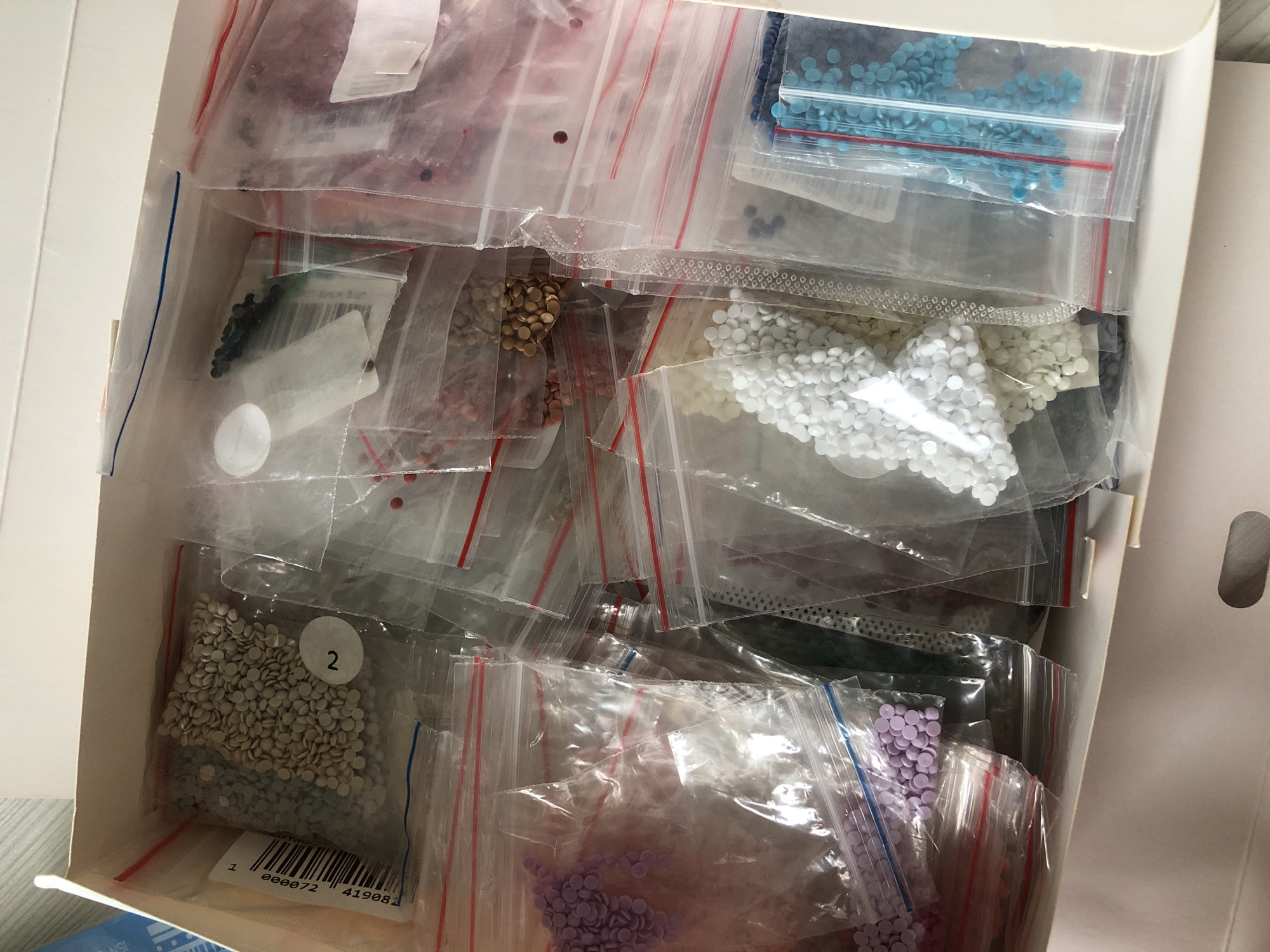 The image shows a white rectangular box filled with numerous small, clear plastic bags. Each bag contains an assortment of colored, round beads, likely for a craft project such as diamond painting. The beads are various colors, including shades of purple, blue, white, gold, and grey. Some bags have labels with numbers or barcodes. The bags are densely packed within the box. The environment appears to be indoors, with a hint of a light-colored surface visible on the left side of the image. The image does not provide any information on the location in the city of Noviy, Russia, or any other details.