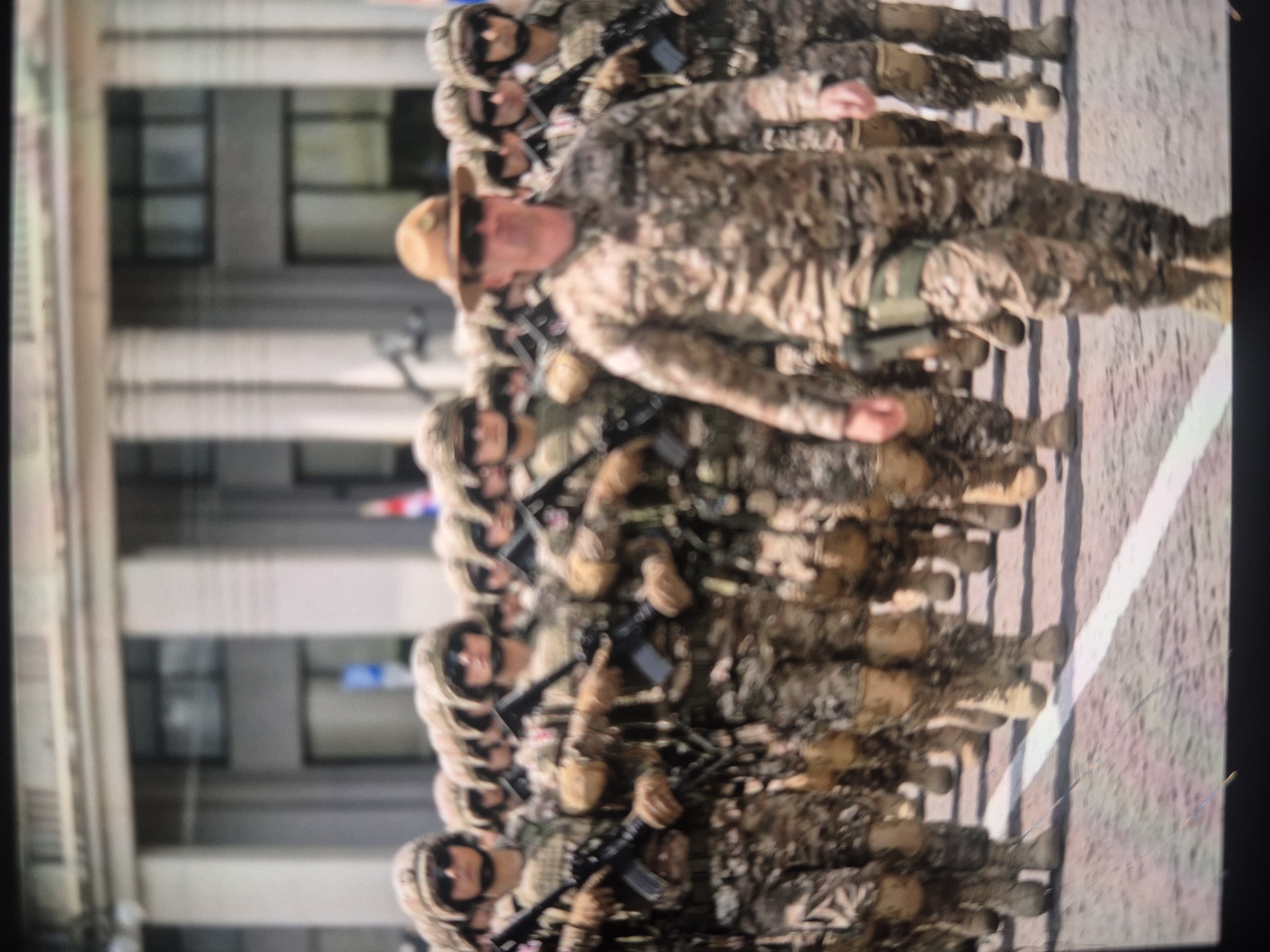 A group of uniformed soldiers marches in formation on a city street near a white painted line. The soldiers are wearing camouflage uniforms, sunglasses, and some have hats. Most are carrying rifles. The background shows a building with classical columns and windows, indicating an urban setting. The scene is likely taking place during a parade or military event, the city is Tbilisi, Georgia.