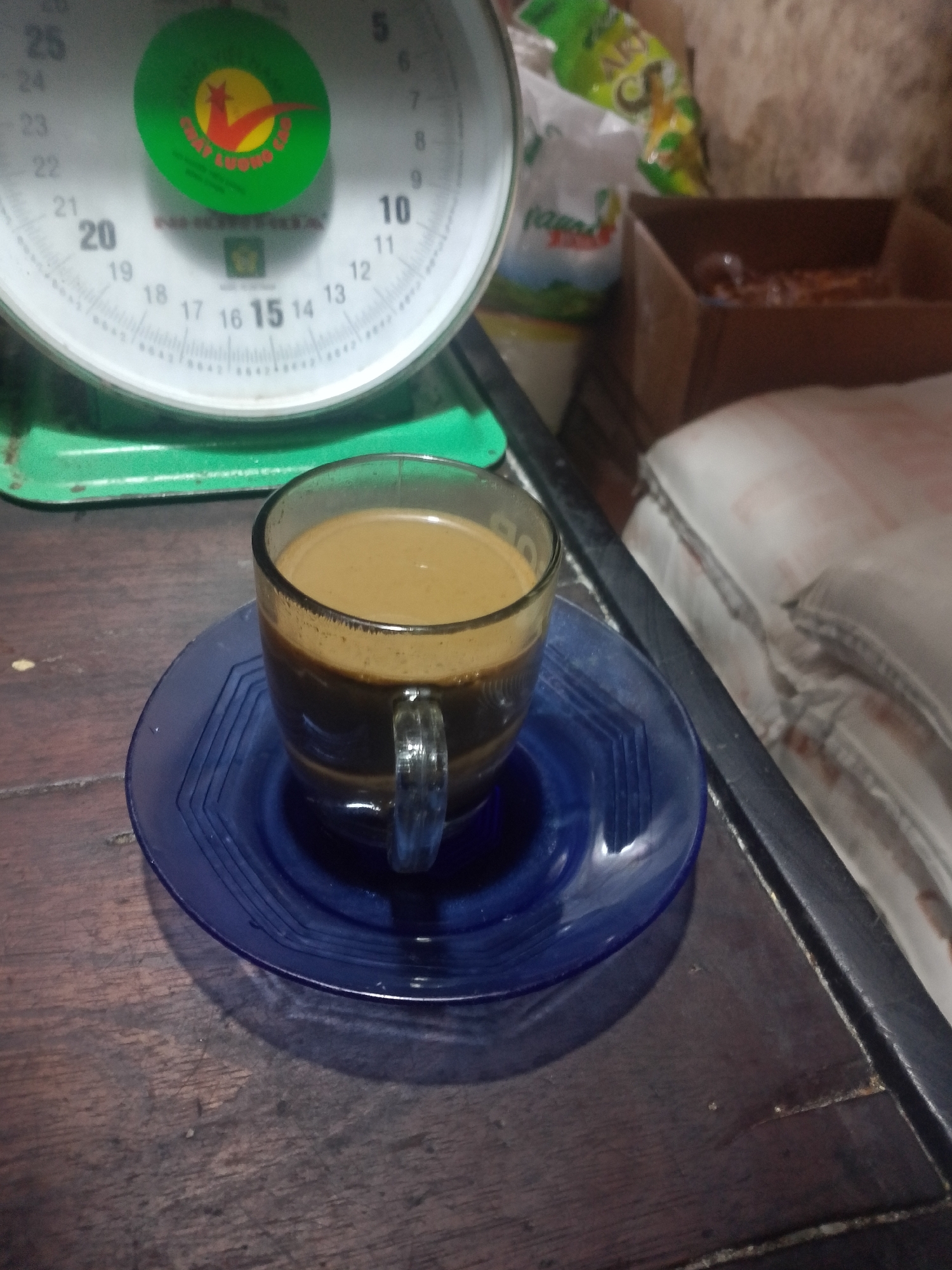 A glass cup of coffee sits on a blue saucer on a dark wooden table. Behind the coffee cup is a green weighing scale with markings and numbers. To the right of the weighing scale are several bags and a cardboard box. The background appears to be a wall. The scene is set indoors, with dim lighting. The image does not provide enough information to determine the location in Palembang, Indonesia.