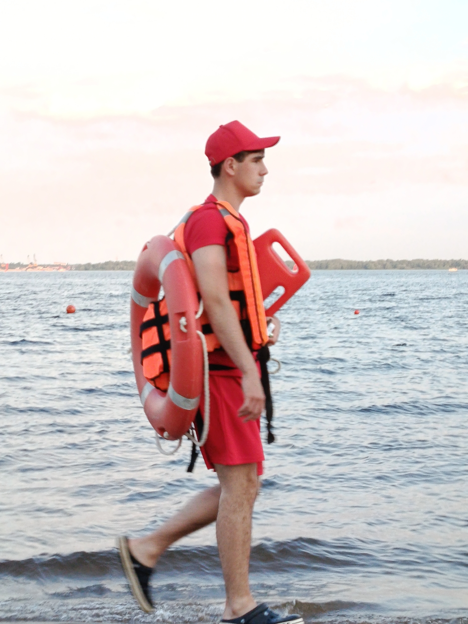 A young man wearing a red cap, red t-shirt, orange life vest, and red shorts walks along the edge of a body of water. He is carrying a red life preserver and a red rescue buoy. The water is choppy, and the sky is overcast with hints of pink and orange. In the background, there is a distant treeline and some structures that appear to be part of a city. There are also several buoys visible in the water.