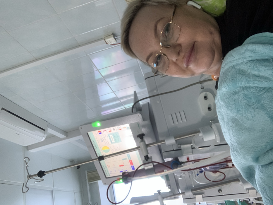 A woman with glasses and a blue robe is positioned near a dialysis machine in what appears to be a medical facility. The machine, displaying medical data on its screen, is connected to tubes. The room has a tiled ceiling and an air conditioning unit. The woman has an earbud in her ear.