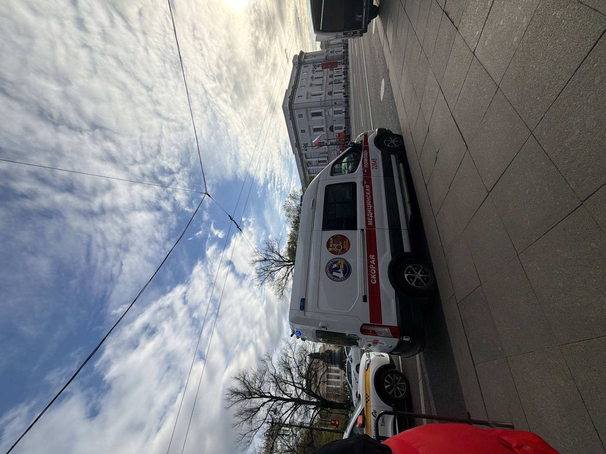 The image shows a street scene in Saint Petersburg, Russia. A white ambulance with red markings is parked on the side of the road. On the side of the vehicle are cyrillic inscriptions. Several more cars can be seen behind the ambulance. A large grey building, likely a government building, is visible in the background. Overhead tram lines are suspended against a backdrop of a cloudy sky. The scene is well-lit, indicating daylight conditions.