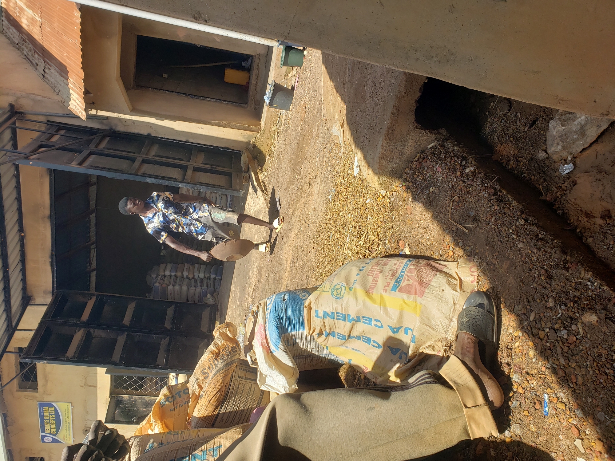 In Jos, Nigeria, a man is seen carrying a round, shallow object as he walks away from an open warehouse filled with stacked bags. The warehouse's large metal doors are open, revealing rows of bagged goods inside. The ground outside the warehouse is dusty and scattered with small stones and debris. In the foreground, a person's legs are visible, wearing brown trousers and sandals, positioned next to several large, labeled bags of cement. To the right, a concrete structure with an opening leads to a dark interior. Two green buckets are placed near the entrance of another building, and a yellow jerrycan is visible inside. A sign on the yellow building reads "VERAZI'S GLOBAL CONCEPTS LTD."