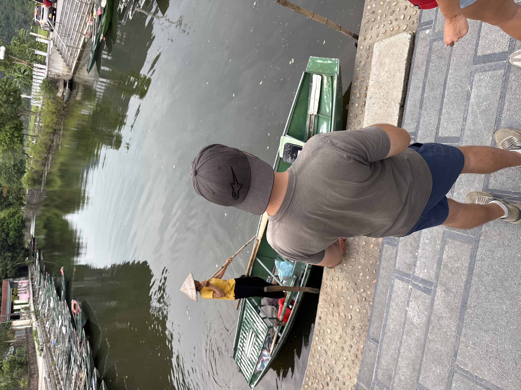 The image depicts a scenic view of a river in Nam Hoa Lư Ward, Vietnam, likely a tourist area. In the foreground, a person wearing a gray t-shirt and a dark baseball cap stands with their back to the camera, looking towards the river. They are positioned on a stone pathway with steps leading down to the water.
The river is calm and reflects the surrounding greenery and overcast sky. Several narrow, green boats are lined up along the riverbank. One boat, closer to the foreground, is occupied by a person wearing a traditional conical hat and a yellow shirt, who is paddling with a long oar. This boat contains various items, including red and blue bags.
The environment is lush with trees and vegetation on the far bank of the river. Buildings and structures are visible in the background, suggesting a developed area. The overall impression is one of a tranquil and picturesque riverside location.