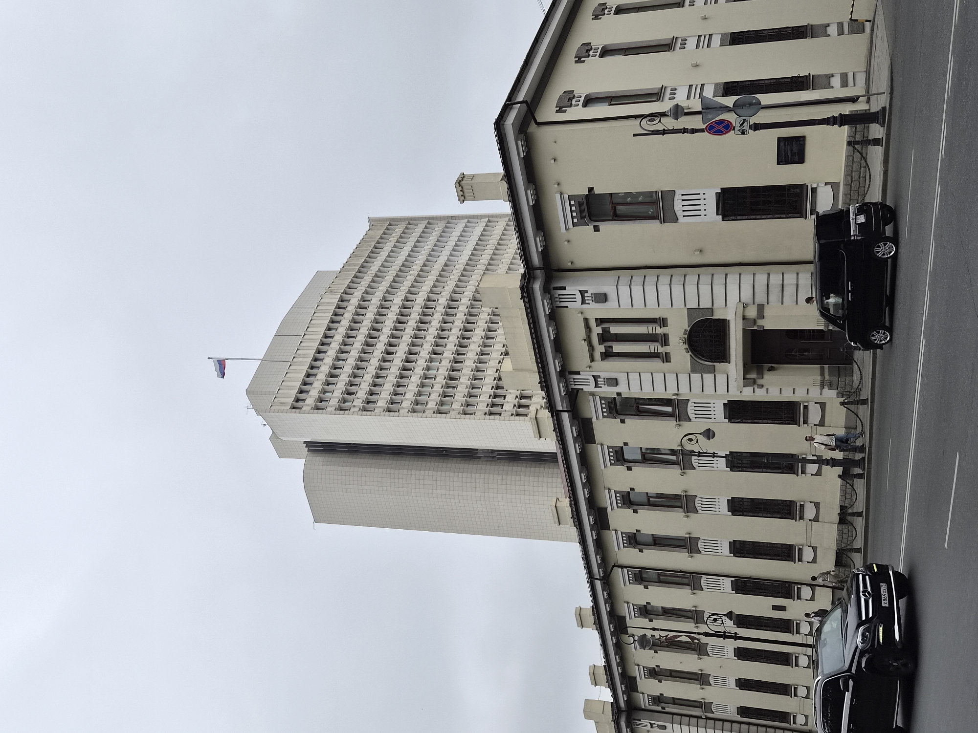 In the city of Vladivostok, Russia, an overcast sky looms over a wide, paved street in front of a pale yellow, two-story building with ornate architectural details and dark-paned windows. A tall, modern, cream-colored building with a grid of small windows stands behind the two-story structure. A Russian flag flies atop the taller building. A black Mercedes SUV is driving on the street, and a black van is parked on the side. Several pedestrians are walking along the sidewalk in front of the yellow building, which is bordered by a low fence.