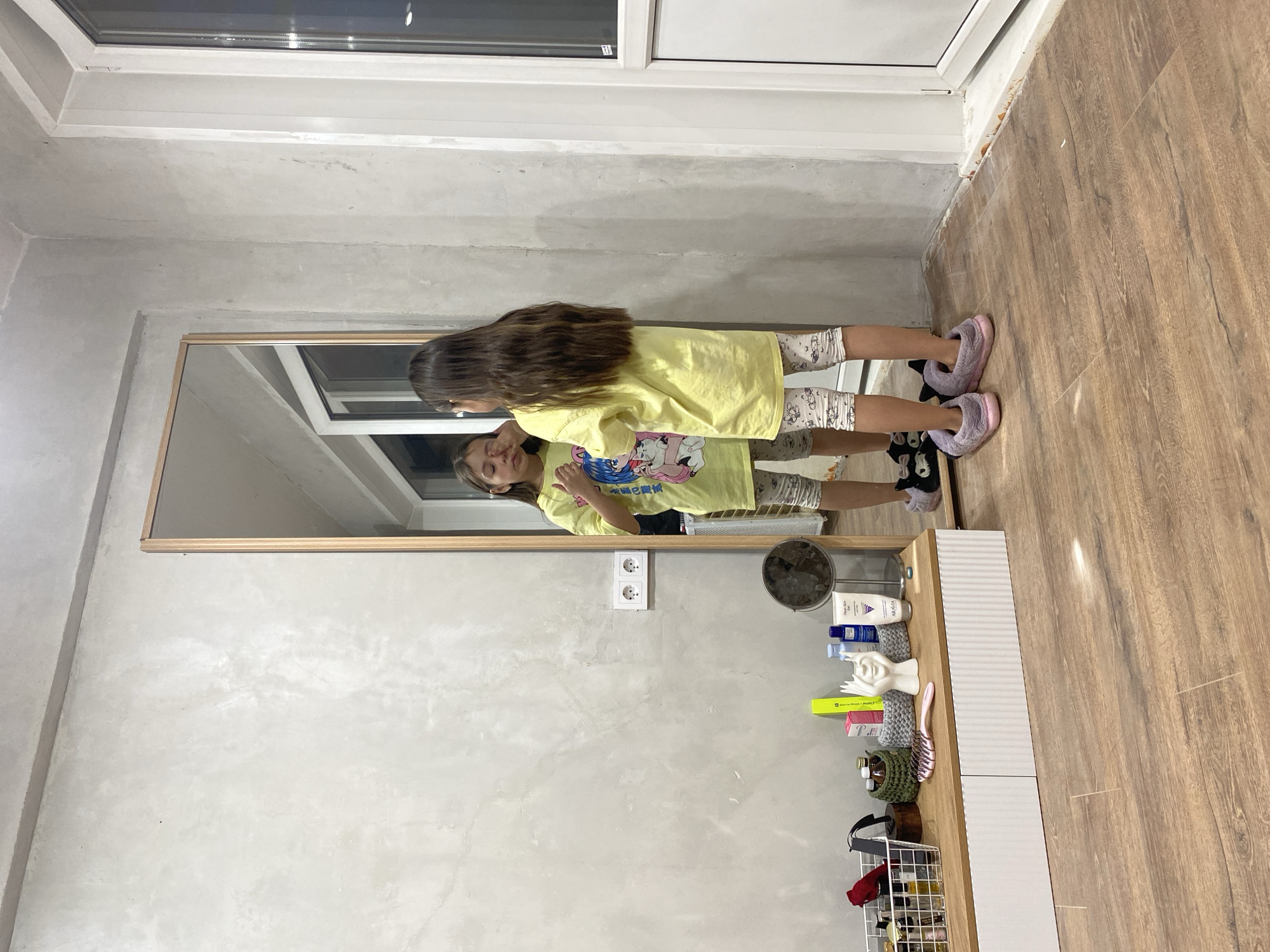 A girl in a yellow t-shirt is standing in front of a mirror. The mirror is hanging on a wall with a gray, textured finish. Next to the mirror is a vanity or shelf unit with various cosmetic and personal care items. A window is above the mirror. The floor is made of light brown wood. The location is possibly Votkinsk, Russia.
