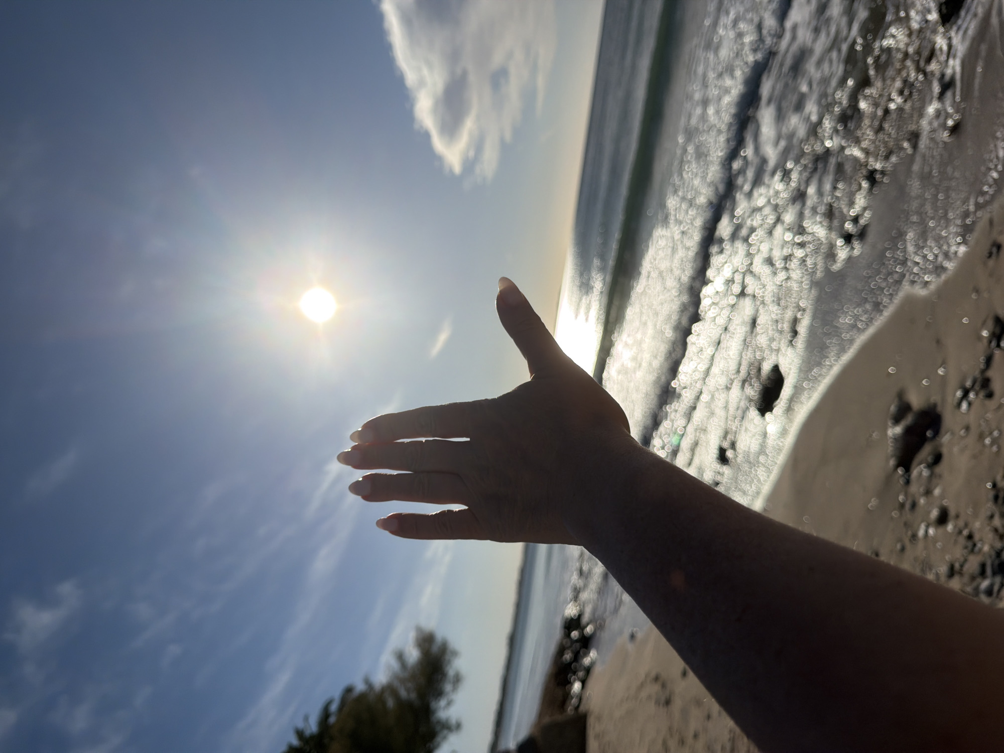 The image captures a bright, sunny coastal scene in Mont-roig del Camp, Spain, viewed from a low angle with a tilted horizon. The primary subject is a human hand, likely female given the manicured nails, extending outwards towards the left, partially silhouetted against the intense sunlight. The hand appears to be interacting with or attempting to shield from the sun.
A brilliant sun, depicted as a strong white light source with a prominent lens flare, dominates the upper left portion of the sky. The sky is predominantly clear blue, with a few faint, scattered clouds.
To the right of the hand, the sea or ocean stretches towards the horizon. The water's surface is highly reflective, featuring numerous sparkling, circular bokeh effects caused by the sun's glare on the rippling waves. The shoreline, composed of sand and possibly small stones, is visible in the lower right corner. In the distant lower left, some dark foliage or trees can be faintly discerned, indicating land.
The time of day is clearly during peak sunshine, likely midday or early afternoon, given the brightness and intensity of the sun. The overall impression is one of a warm, sunny day at the beach, evoking feelings of relaxation and enjoyment of the natural environment. There is no visible text in the image.