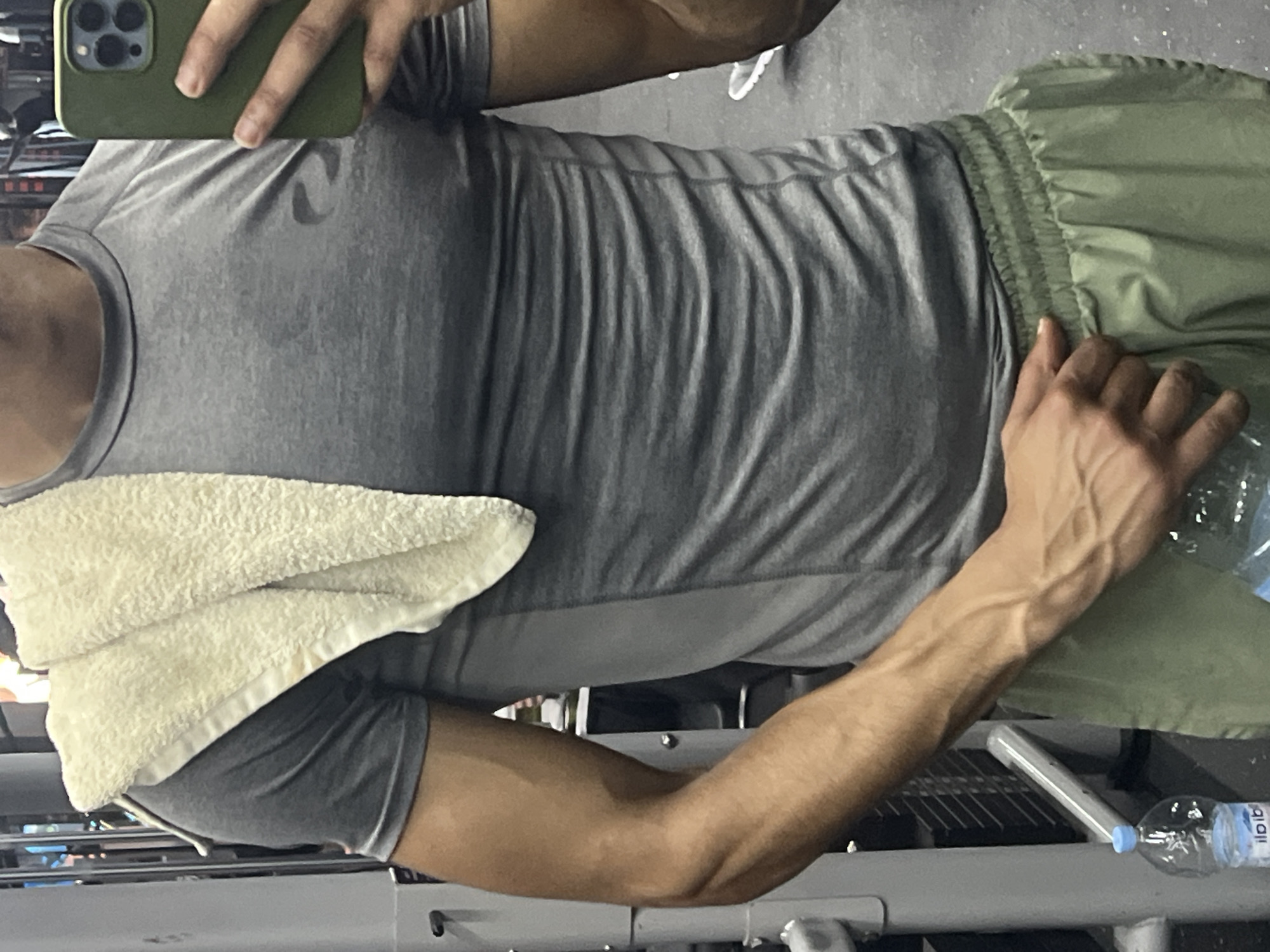 The image captures a waist-up view of a person, seemingly taking a mirror selfie in an indoor gym setting. The individual is wearing a grey athletic t-shirt, which appears to have a subtle Nike swoosh logo on the chest, and light green athletic shorts with a ruched elastic waistband. A cream-colored towel is draped over their left shoulder and chest. The person's left hand, holding a dark green-cased smartphone with a triple camera array, is visible at the top of the frame. Their right arm, extended downwards, displays a muscular forearm with prominent veins. They are holding a clear plastic water bottle with a blue cap in their right hand. The background features metallic frames and bars consistent with gym equipment. The water bottle's label clearly shows the text "ilpib".