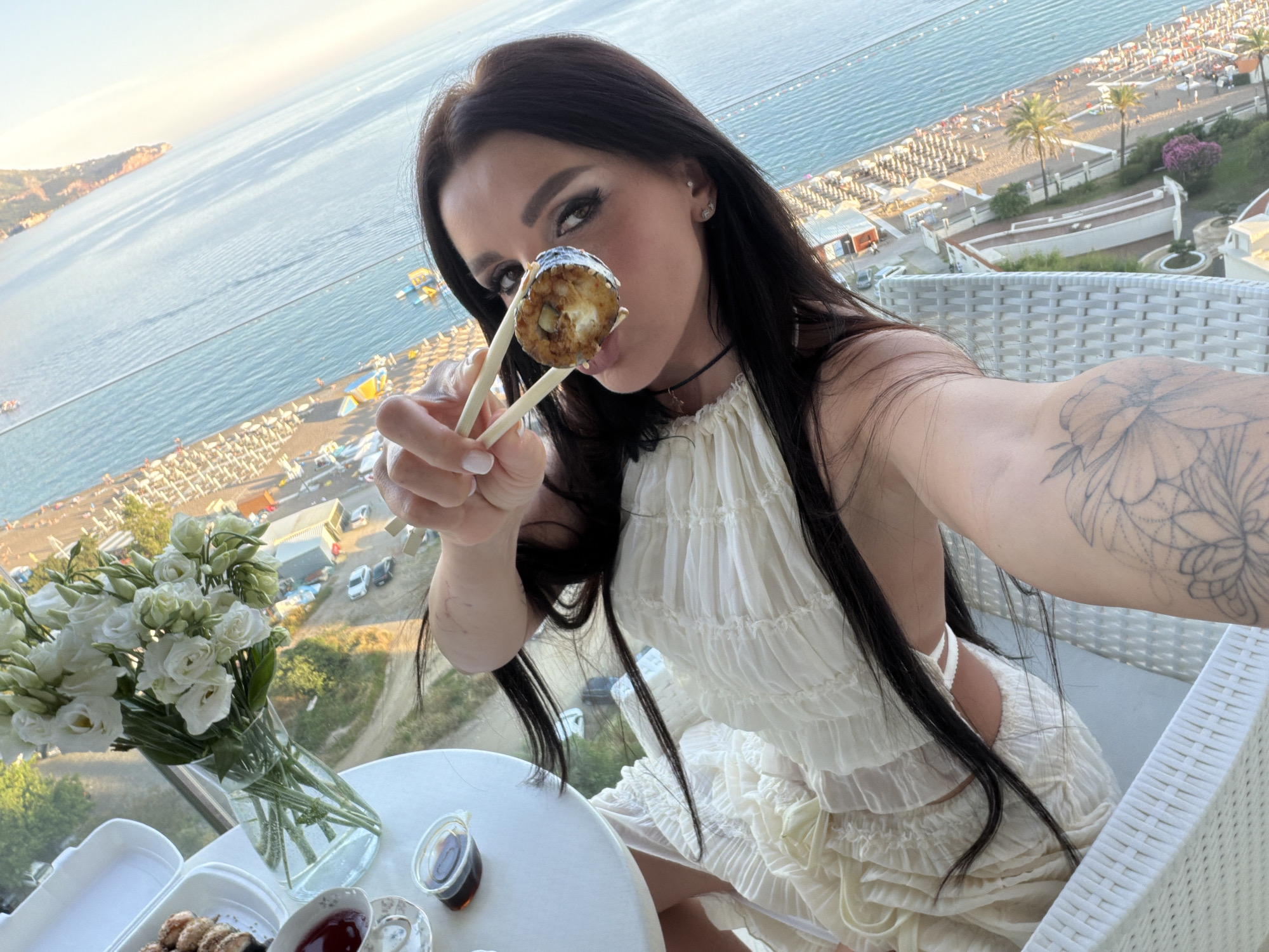 A woman with long dark hair and heavy eye makeup is holding a piece of sushi with chopsticks, bringing it towards her mouth. She is seated at a table on a balcony overlooking a beach and the sea. The table has a vase of white flowers, a container of sushi, and small dishes of sauce. The woman is wearing a light-colored, ruffled dress. The backdrop shows a beach with numerous lounge chairs, palm trees, and the ocean. The woman has a tattoo on her left arm. The scene appears to be in Boreti, Montenegro.
