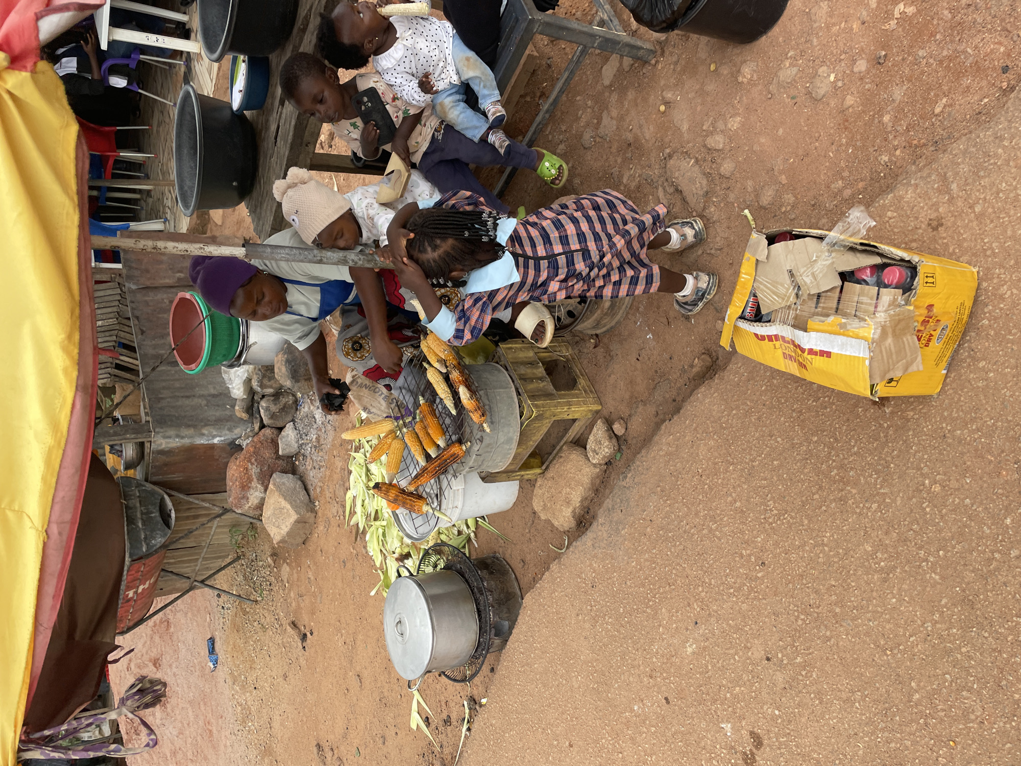 The image depicts an outdoor scene in Jos, Nigeria, characterized by a makeshift food stall under a yellow and red awning. A woman, dressed in a blue apron over patterned pants and a purple headscarf, is attending to a grill laden with corn. She appears to be cooking and interacting with a group of children gathered around.
Several children of varying ages are present. Two young children are positioned close to the woman, one wearing a light-colored knitted hat and the other in a checkered dress. Further to the right, two older children are seated on a bench; one is holding a black object, possibly a phone, and the other is eating corn. Another child, wearing a white polka-dot shirt, is also seated on the bench, holding a piece of corn. A young girl in a plaid dress stands near the grill, her hands on her head.
The environment is rustic, with sandy, dirt ground. In the background, there are various containers, including large black basins, a green bucket, and metal pots. Some of the corn husks are scattered around. To the lower right of the image, a yellow cardboard box containing bottles is visible, partially opened and taped. The overall impression is one of a busy, informal marketplace scene.