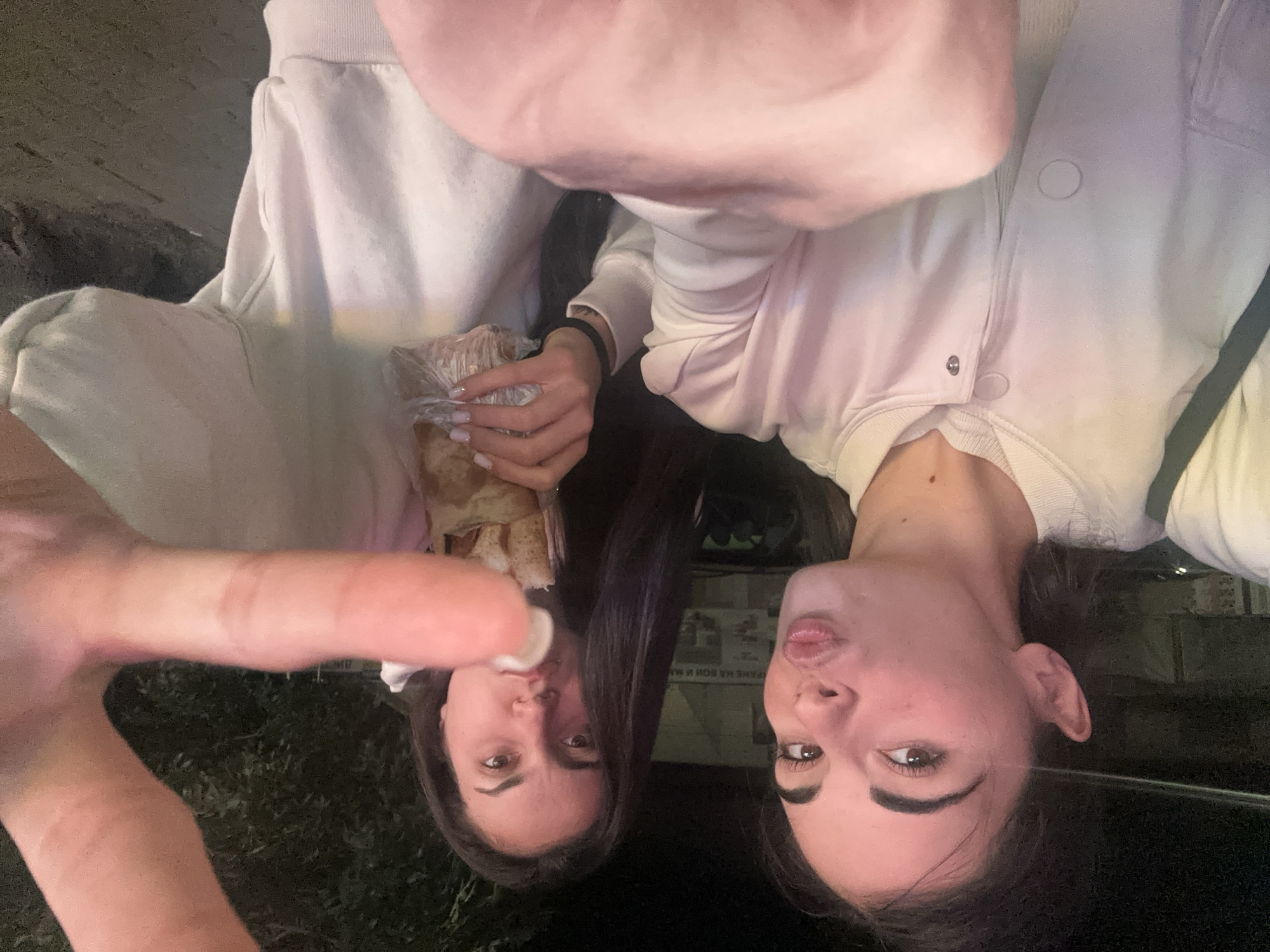 Two women are captured in a selfie at night in an outdoor urban environment. The woman on the left, in the foreground, is making a kissing gesture with her lips pursed. She has dark hair and is wearing a light-colored jacket over a white top. Her right hand is visible in the lower right portion of the frame. The woman on the right is holding a food item wrapped in plastic and paper, and appears to be taking a bite. She has dark hair and is wearing a white sweatshirt. Her left hand is out of frame. Behind the women, a building with some signage is visible, along with trees and foliage in the background. The lighting suggests it is nighttime, with artificial lights illuminating the scene. The ground appears to be a paved or dirt surface.