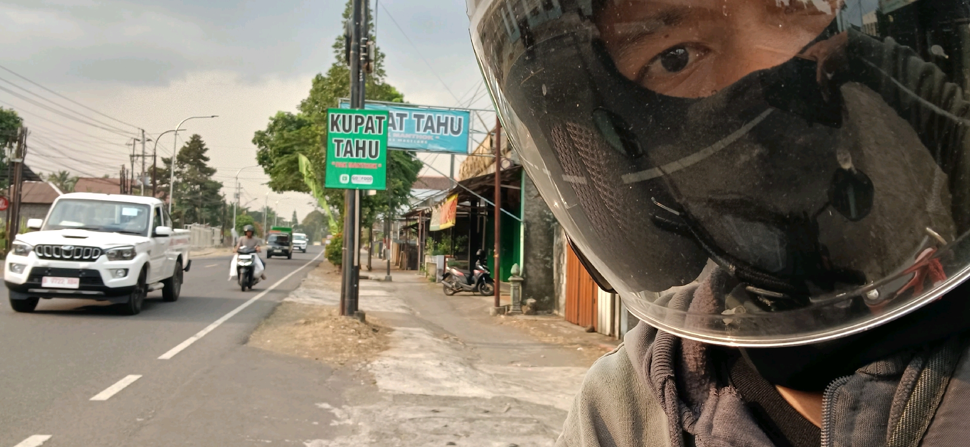 A first-person perspective, likely taken by someone on a motorcycle, shows a street scene in Badran, Indonesia. The foreground is dominated by the rider's helmet and face, with their right eye visible through the visor. The helmet is black and reflects the surrounding environment, including the road, buildings, and other vehicles.
The street is paved with asphalt and has a white dashed line marking the lane. To the left, a white pickup truck is driving in the same direction as the viewer. Further down the road, a person on a motorcycle is visible, also traveling in the same direction. On the right side of the road, there are shops and buildings lining the street. One shop has a green sign that reads "KUPAT TAHU TA HU" and another sign above it says "GO FOOD". A few motorcycles are parked in front of the shops.
The environment appears to be a mix of urban and suburban, with trees and power lines visible in the background. The sky is overcast, suggesting it might be a cloudy day. The overall scene captures a typical street view in Indonesia, with its characteristic modes of transportation and commercial establishments.