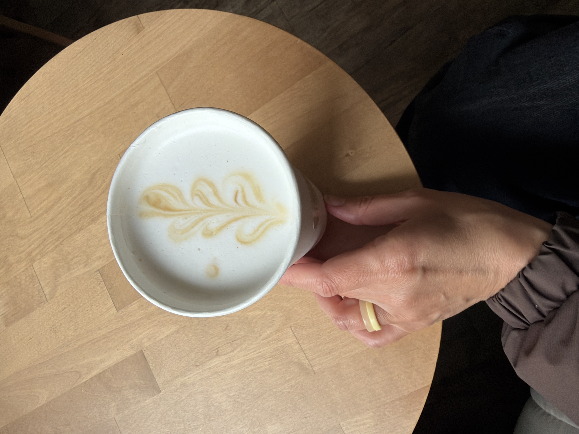 A person's hand is holding a cup of latte art on a light brown, round wooden table. The person wears a light yellow ring and part of their dark blue jeans and a brown jacket are visible. The table appears to be indoors. The image does not provide any specific information to verify the location in Chaikovsky, Russia.