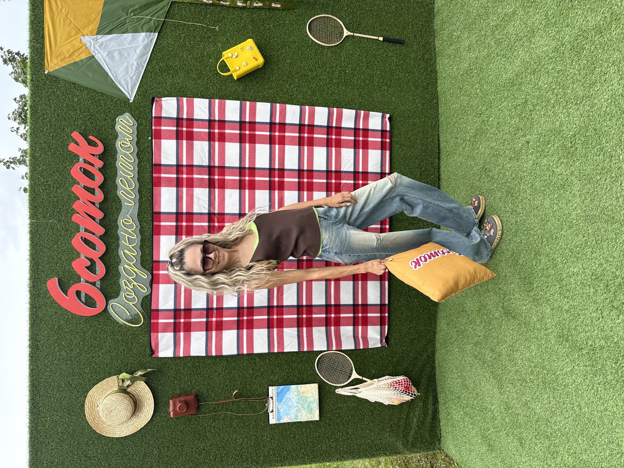 The image shows a woman standing on artificial grass in front of a green wall decorated with various objects. The wall features a large red and white checkered blanket, a straw hat with a green ribbon, a vintage brown camera, a map on a clipboard, a tennis racket, and a string bag with fruit. To the right, there is a kite in green and yellow, a yellow purse adorned with charms, and another tennis racket. Red neon lettering above the blanket reads "6 соток" (6 hundred square meters), and yellow neon lettering below it reads "Создано летом" (Created in summer). The woman is wearing a brown tank top with a neon green trim, distressed blue jeans, and platform shoes. She is holding an orange pillow with red lettering. The overall scene appears to be a promotional setup, possibly for a summer event or a dacha-related theme, located in the city of Zhukovo, Russia, as indicated by the lettering.