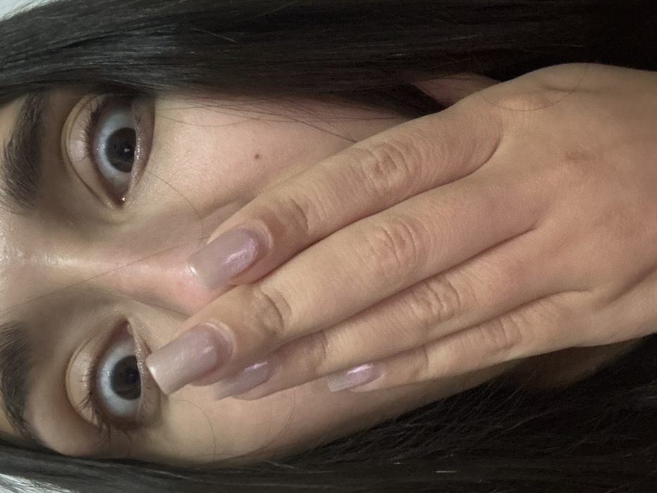 A close-up, slightly tilted view of a person's face, partially obscured by their hand. The person has wide, expressive eyes and dark hair. Their hand covers the lower portion of their face, with long, squared-off nails that have a subtle, iridescent polish. The skin is fair. A small mole is visible near the nose. The lighting is soft, emphasizing the textures of the skin and hair.