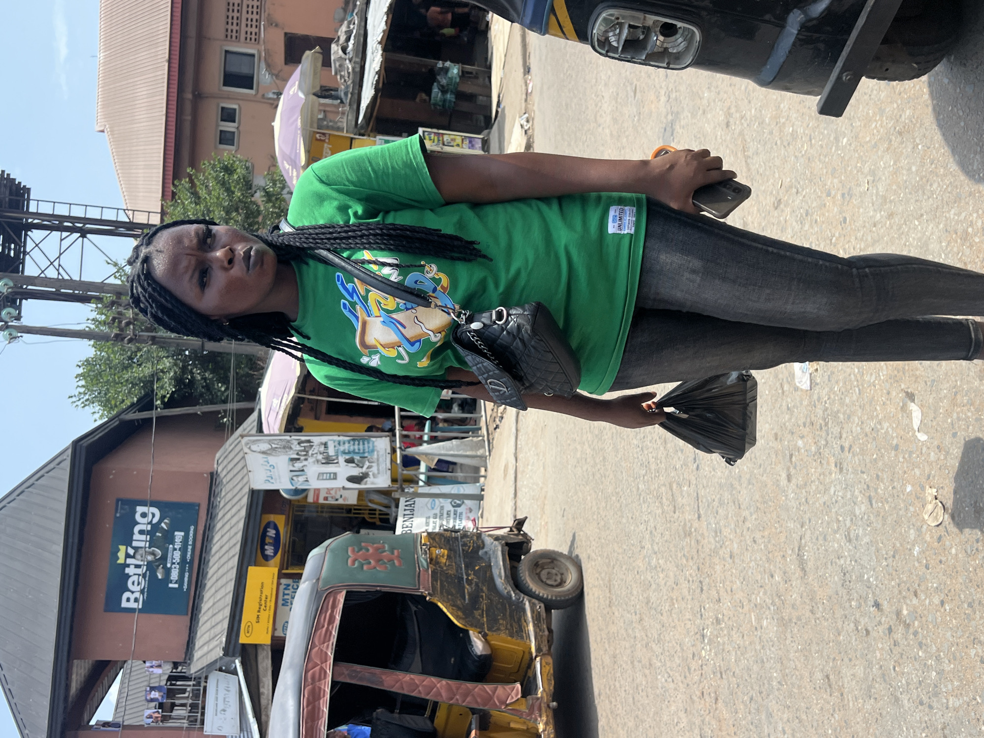 The image shows a woman in Onitsha, Nigeria, standing on a street. She has braided hair, wears a green t-shirt, dark jeans, and a black purse. She is holding a black plastic bag in one hand and a phone in the other. Behind her is a yellow auto-rickshaw, with "BENIJAN" visible on a sign nearby. A building with a "BetKing" sign is also visible in the background, along with other shops. The sky is clear, suggesting it is daytime.