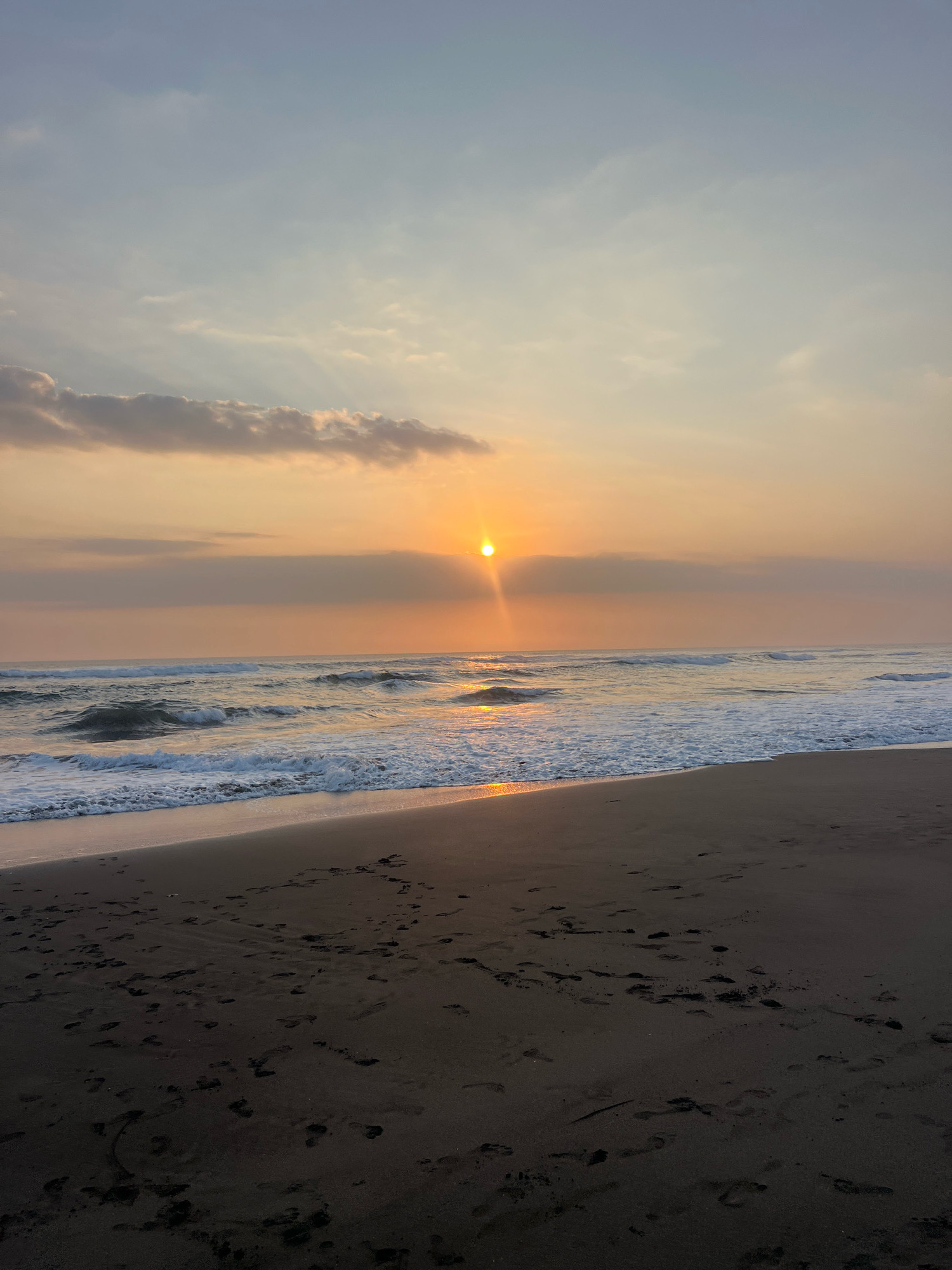 A serene beach scene unfolds at sunset, with the sun appearing as a bright orb just above the horizon, casting warm, golden light across the sky and water. The sky transitions from a soft blue at the top to a gradient of peach and orange hues near the horizon, with scattered clouds tinged with the sunset colors. Rays of light emanate from the sun, creating a dramatic effect.
The ocean is calm, with gentle waves rolling towards the shore, their crests marked by white foam. The water reflects the colors of the sunset, creating a shimmering path of light on its surface. A sandy beach occupies the foreground, appearing dark and textured with numerous footprints imprinted on its surface. The footprints suggest recent human activity on the beach.
The overall atmosphere is tranquil and picturesque, evoking a sense of peace and natural beauty. It is unlikely that this specific beach is located within the city of Kediri, Indonesia, as Kediri is an inland city, not a coastal one. This scene is characteristic of a coastal environment rather than an urban setting.