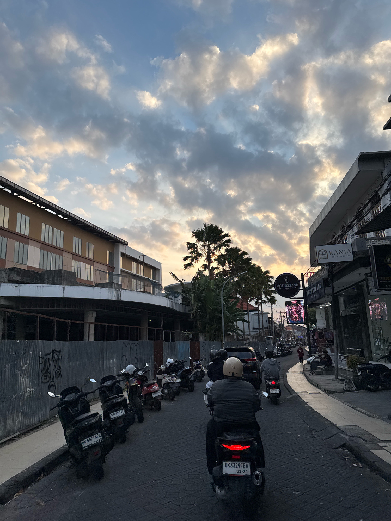 The image depicts a street scene in Kuta, Indonesia, during dusk. The sky is filled with dramatic clouds illuminated by the setting sun. On the left side of the street, a multi-story building with a tan facade and reflective windows is under construction, fenced off with corrugated metal. On the right side, a row of shops and businesses lines the street, with prominent signage for "KANIA ROOMS & SUITES" and "MOTHERLAND".
The street is busy with traffic, primarily consisting of scooters. Several scooters are parked along the side of the road, while others are in motion. In the foreground, a person wearing a beige helmet is riding a black scooter. Further down the street, other individuals are visible on scooters and walking. Palm trees are scattered throughout the scene, adding to the tropical atmosphere. The road is paved with cobblestones. The overall impression is one of a lively, urban environment at the end of the day.