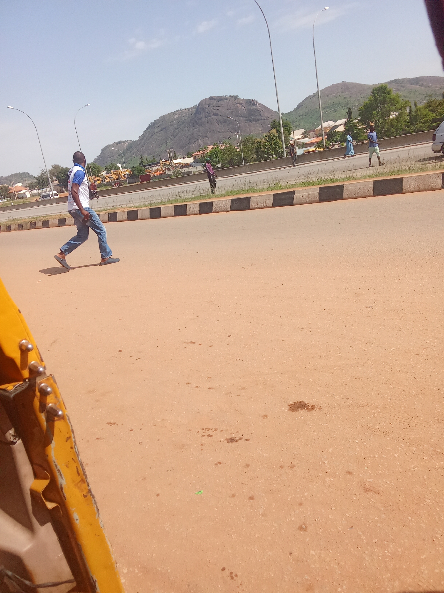 A man in a white t-shirt with blue trim and blue jeans walks across a paved road. He is on the right side of the frame, moving towards the left. In the background, a highway with streetlights runs parallel to a rocky, green mountain range. Several buildings and trees are visible between the road and the mountains. Other pedestrians and a vehicle are also present in the background. The road surface is a light brown color and appears to be asphalt.