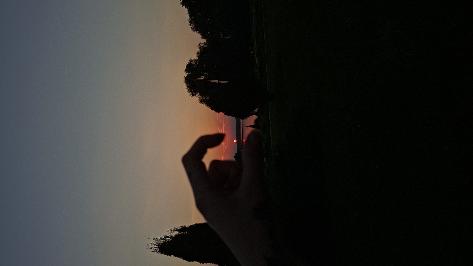 The image captures a silhouette of a hand framing a sunset over a body of water. The sun, a small, vibrant red orb, is partially visible just above the horizon, casting a reflection on the water. The sky transitions from a deep blue at the top to a warm orange and pink near the horizon. Silhouetted trees and vegetation form the background, creating a dark, natural landscape. The foreground is dominated by the dark silhouette of a person's hand, with the thumb and index finger forming a circular shape around the setting sun. This scene appears to be located near the city of Myadzyel, Belarus, given the natural surroundings and the common landscape features found in the region during sunset.