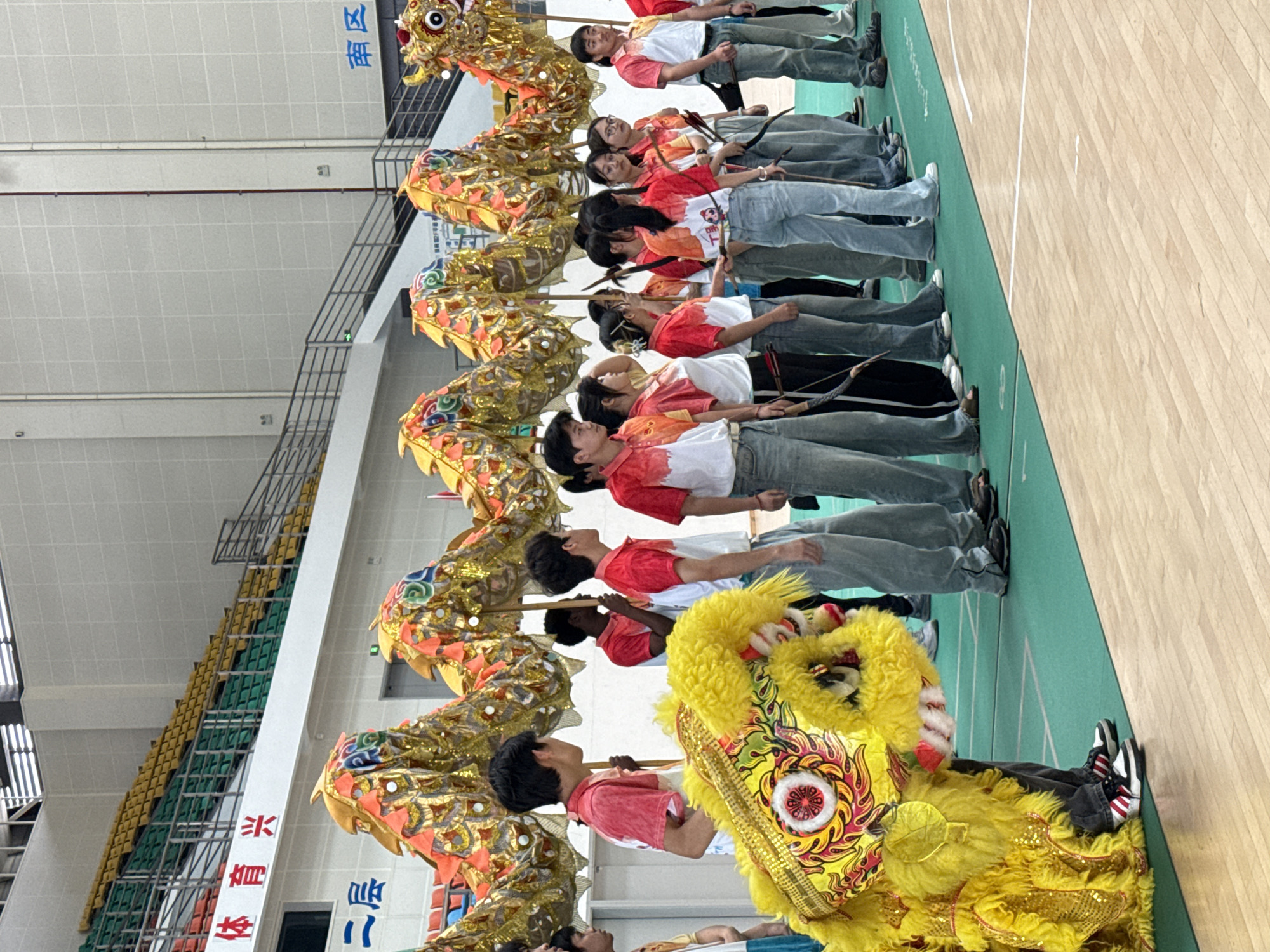 In a large indoor space in Jiangjin, China, a group of approximately 10 people are lined up, dressed in red and white shirts and jeans. They are positioned on a green and wooden gymnasium floor. Most hold long, thin sticks. Above them is a long, elaborate gold-colored dragon costume, likely for a dragon dance performance. At the front of the line is a large yellow lion costume, common in Chinese lion dances. The background includes rows of empty stadium-style seating, a high ceiling with lighting, and banners with Chinese characters.