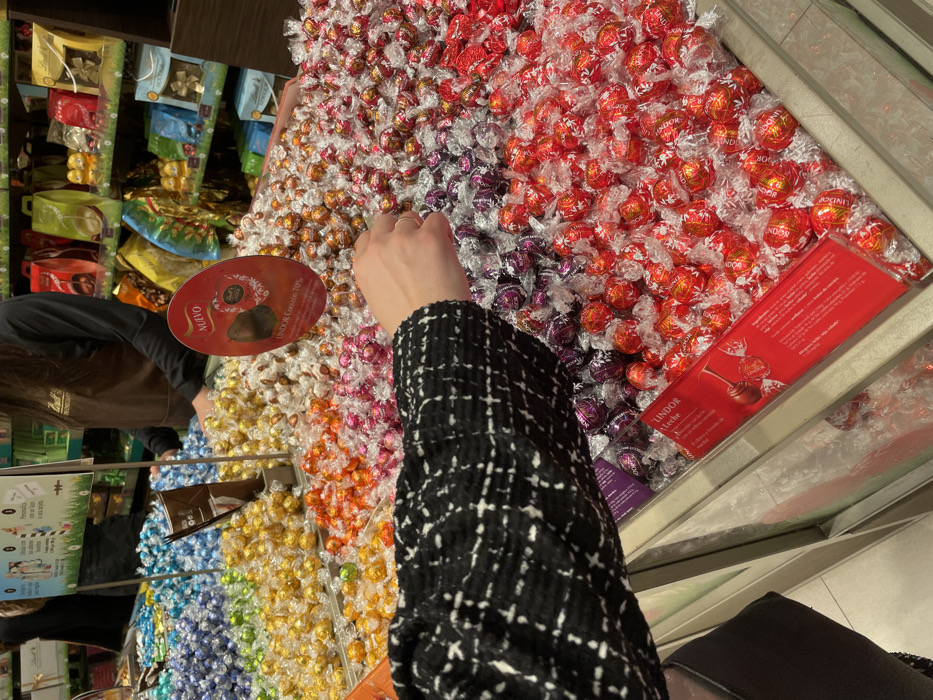 The scene depicts an interior view of a Lindt chocolate store or concession located in Marratxí, Spain. In the foreground, the left arm of an individual, wearing a black and white patterned tweed-style jacket, extends into a large, multi-compartment display bin filled with bulk, individually wrapped Lindor chocolates. The hand is positioned above sections containing red and purple foil-wrapped truffles.
The extensive display bins showcase a spectrum of Lindor chocolate varieties, color-coded by wrapper, including blue, green, yellow, orange, pink, purple, red, and silver/white. A red oval-shaped sign, centrally placed above the bins, reads "NUEVO" (New) and identifies "LINDOR Corazón 70%." A rectangular red sign at the bottom right of the display labels a section "LINDOR Leche" (Lindor Milk). A brown paper scoop or bag holder is visible within one of the central bins.
In the background, a store employee, partially visible, stands behind a counter. This individual wears a brown apron featuring the Lindt logo. Behind the employee, shelving units are stocked with various packaged chocolate products, gift bags, and boxes, indicative of a retail setting. The area is brightly illuminated, and the flooring appears to be light-colored tiled material.
