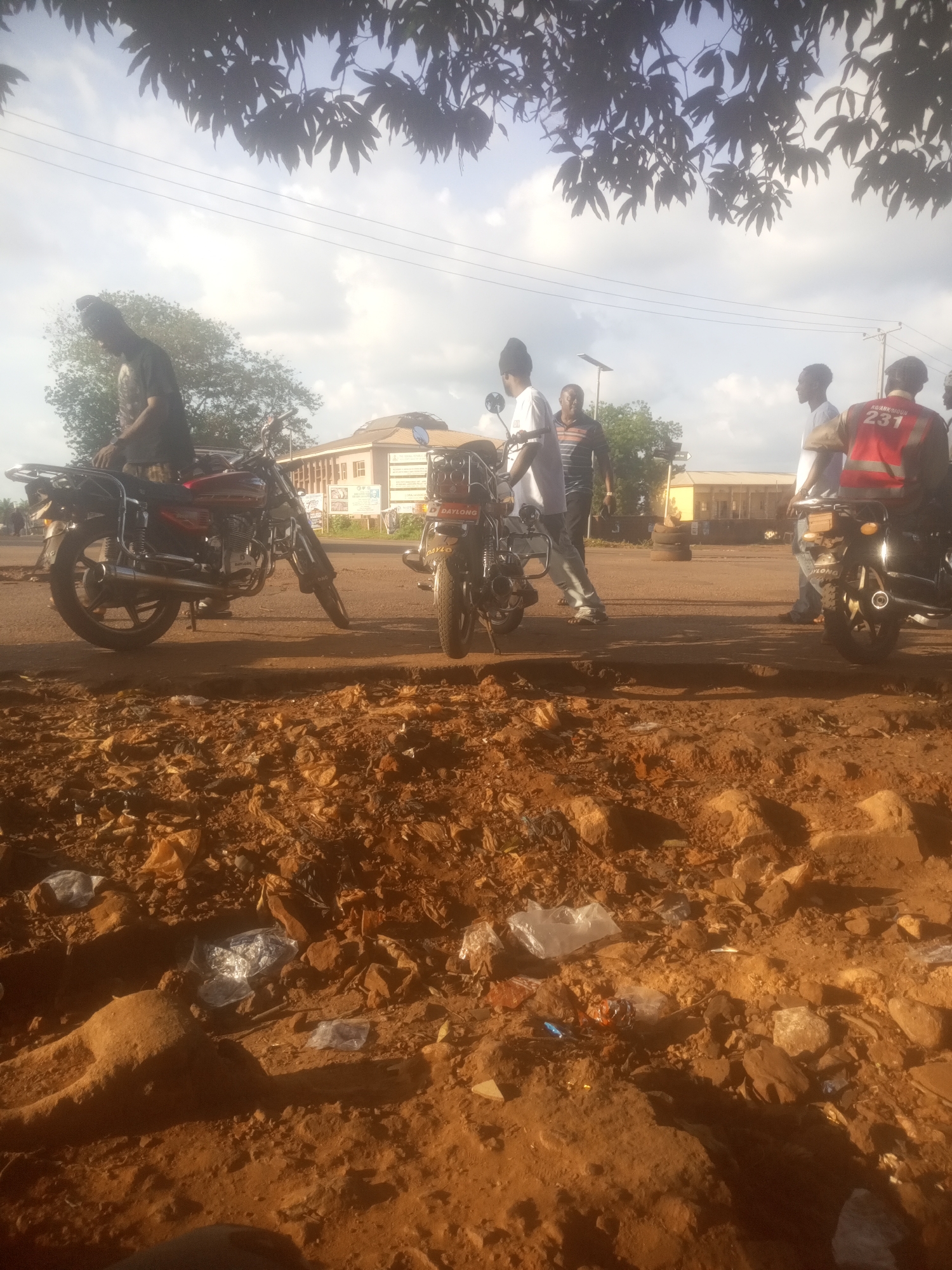 In Ankpa, Nigeria, on a dusty, sunlit day, several individuals are gathered around motorcycles. The foreground is dominated by a rough, reddish-brown dirt surface littered with discarded plastic bags and rocks. In the midground, a red and black motorcycle is parked on the asphalt road, with a person in a dark t-shirt and headwear standing beside it. Further down the road, other individuals are seen near two more motorcycles. One person wears a red vest with the number "231" and "AKAMKONUM" printed on it. In the background, a light-colored building with a curved roof stands against a bright, cloudy sky. A few trees are visible, with their dark leaves silhouetted against the sky. Power lines are strung across the sky.