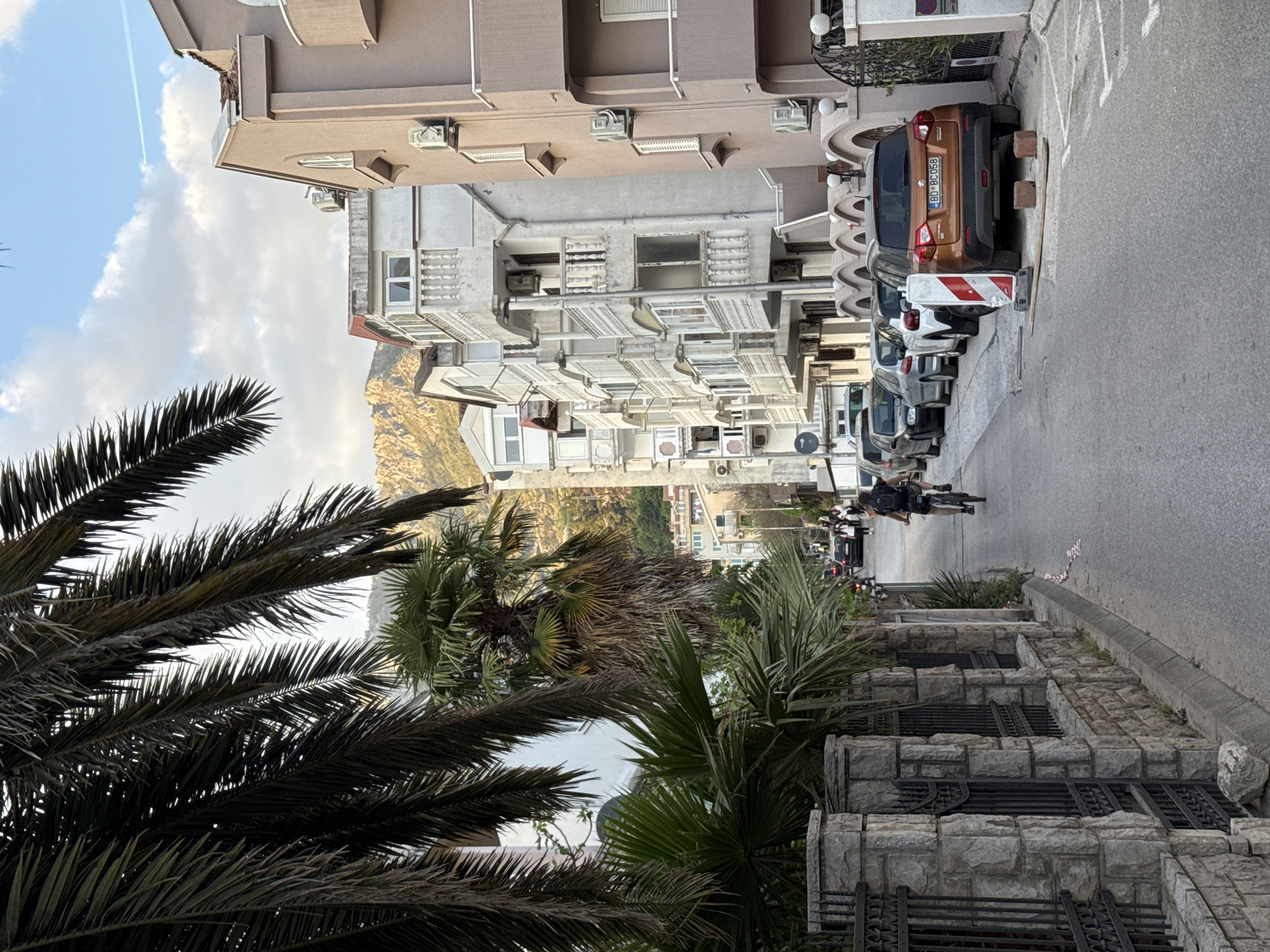 An eye-level medium shot looking up a street in Budva, Montenegro. In the foreground, palm trees obscure parts of the view. A grey stone wall sits on the right, with metalwork details.
The street ascends, with cars parked on the right side. Buildings with many windows and air conditioning units line both sides of the street. A person on a bicycle is visible further up the street. In the distance, a mountain range is visible. The sky is light blue with some clouds.