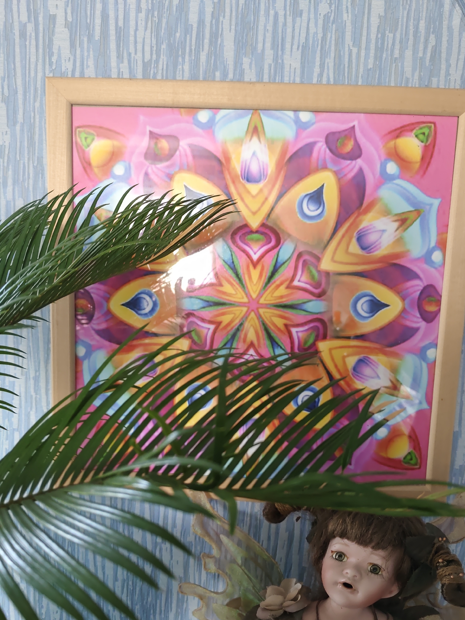 An indoor scene features a framed colorful mandala art piece hanging on a blue textured wallpaper. In the foreground, a potted palm plant's green fronds partially obscure the artwork. Below the artwork, a porcelain doll with green eyes and brown hair is visible, adorned with decorative elements. The overall setting appears to be a room in Rostov-on-Don, Russia, though specific location details within the city are not discernible from the image.