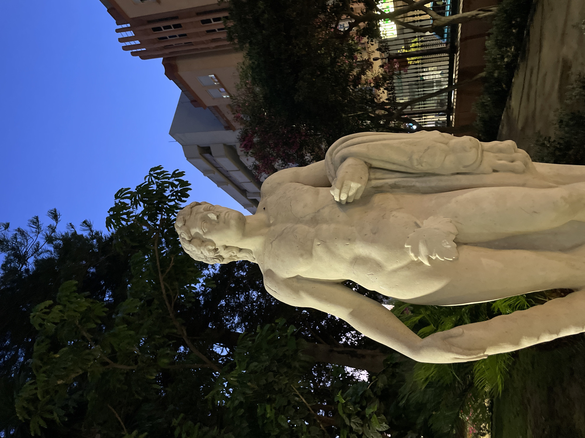 The image shows a full-body, life-size, light-colored stone statue of a male figure. The statue is positioned outdoors in what appears to be a public space at dusk or early evening. The figure is nude except for a fig leaf covering its genitals and a draped cloth held in its right hand. The statue has a muscular build, curly hair, and is looking slightly to its right.
Behind the statue, there are trees with lush green foliage. Further in the background, several multi-story buildings are visible, with some windows illuminated, suggesting urban surroundings. The sky is a clear, deep blue, characteristic of twilight. A black metal fence with ornamental posts runs horizontally in the mid-ground. The ground in front of the statue is paved, with some scattered vegetation and dimly lit areas, indicating it is likely a park or plaza.
The statue is a prominent feature in the foreground, illuminated by an unseen light source that highlights its contours. The overall impression is of a classical sculpture set against a backdrop of a Spanish city. This statue is identified as "El Niño de la Bola" and is located in the Plaza de la Constitución in La Línea de la Concepción, Spain.