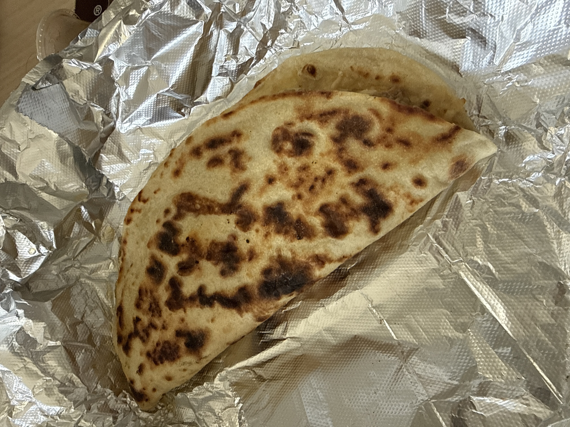 The image shows a folded flatbread, likely a quesadilla or similar item, with browned spots on its surface. It rests on a sheet of crumpled aluminum foil. The image does not provide enough information to determine the location in the city of New York, United States of America. There are no people visible in the image. The object is a folded flatbread on aluminum foil. There are no actions or interactions visible. The environment consists of the foil and the surface on which it rests.