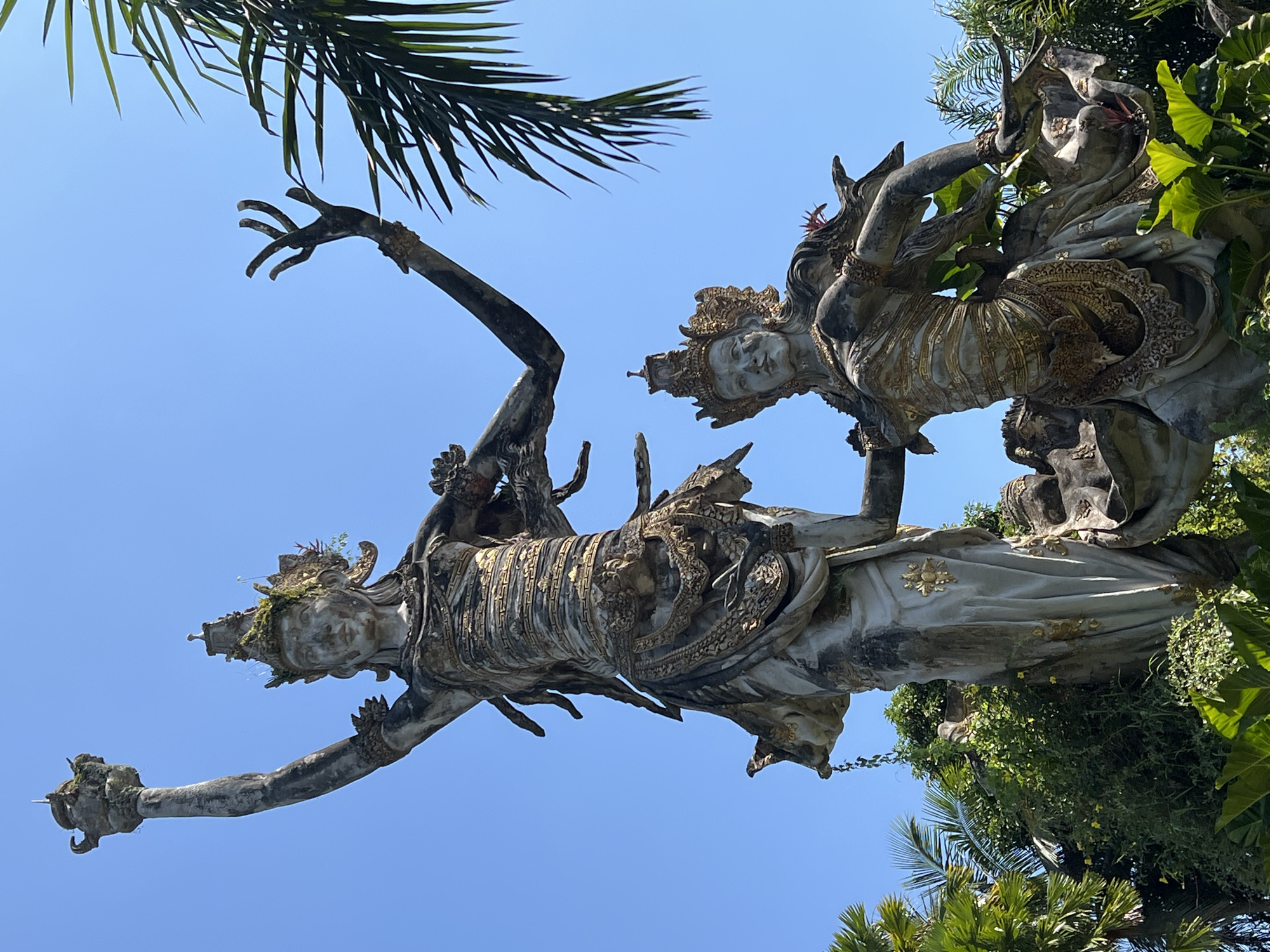 The image captures a view of a Balinese sculpture in Kedewatan, Indonesia, against a clear blue sky. The sculpture depicts two human-like figures adorned with intricate detailing and crowns. The statues are positioned amongst lush foliage, with palm fronds visible both above and on the right side of the image. The statues are a grey, stone-like material with gold detailing. The overall orientation of the image suggests it was taken from below, looking upwards towards the statues and the sky.