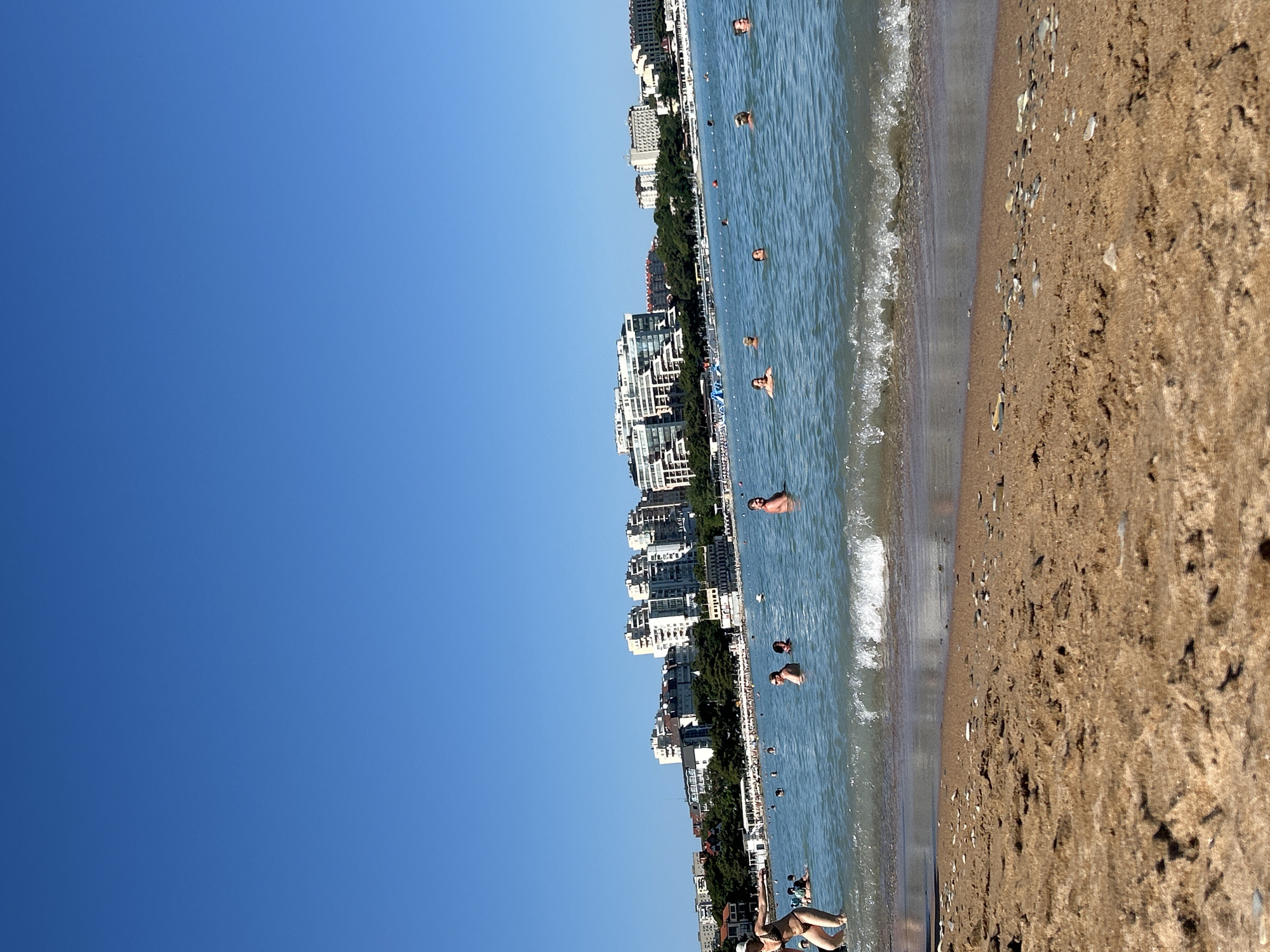 The image depicts a bustling beach scene on a clear, sunny day, likely in Gelendzhik, Russia, given the distinctive architecture of the buildings visible in the background. The foreground is dominated by a sandy beach, with some scattered pebbles and the gentle lapping of waves.
In the shallow water, numerous people are enjoying a swim. Towards the left of the frame, a woman in a bikini and a white sun hat is wading in the water, facing away from the camera and extending her left arm. Further into the water, several individuals are visible, some with their heads above the surface, engaged in swimming or relaxing. A small wave is breaking near the shore, adding to the dynamic nature of the scene.
The middle ground features a more crowded area of the sea with many more swimmers. The background reveals a prominent cityscape characterized by a series of multi-story buildings, many of which appear to be hotels or apartment complexes, lining the coastline. These structures exhibit modern architectural styles with balconies and light-colored facades. Lush green trees are interspersed between the buildings and along the beachfront. The clear blue sky stretches across the upper portion of the image, suggesting fair weather. The overall impression is one of a popular tourist destination during peak season.