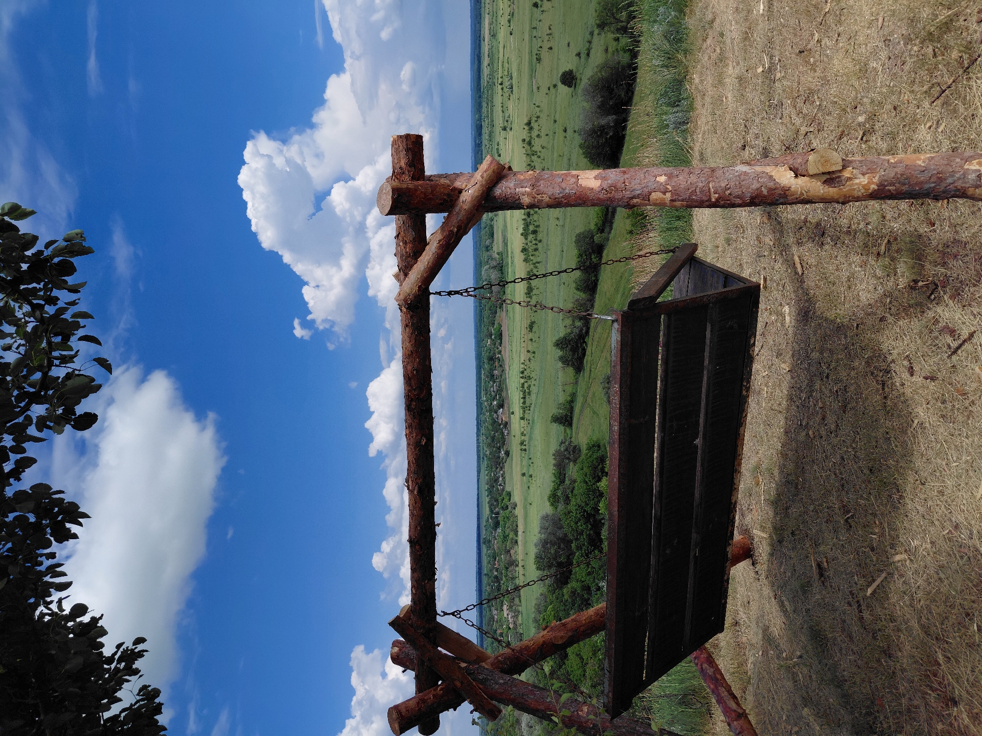 The image depicts a wooden swing set placed on a grassy hill, overlooking a sprawling rural landscape under a bright blue sky with scattered white clouds. The swing set is constructed from rough, natural logs, with a dark wooden seat suspended by chains. In the distance, a valley unfolds, dotted with scattered houses and lush green fields and trees. The foreground is characterized by dry, yellowed grass and scattered twigs. The scene appears to be in Ustyvytsia, Ukraine, suggesting an outdoor recreational area or viewpoint. There are no people visible in the image.