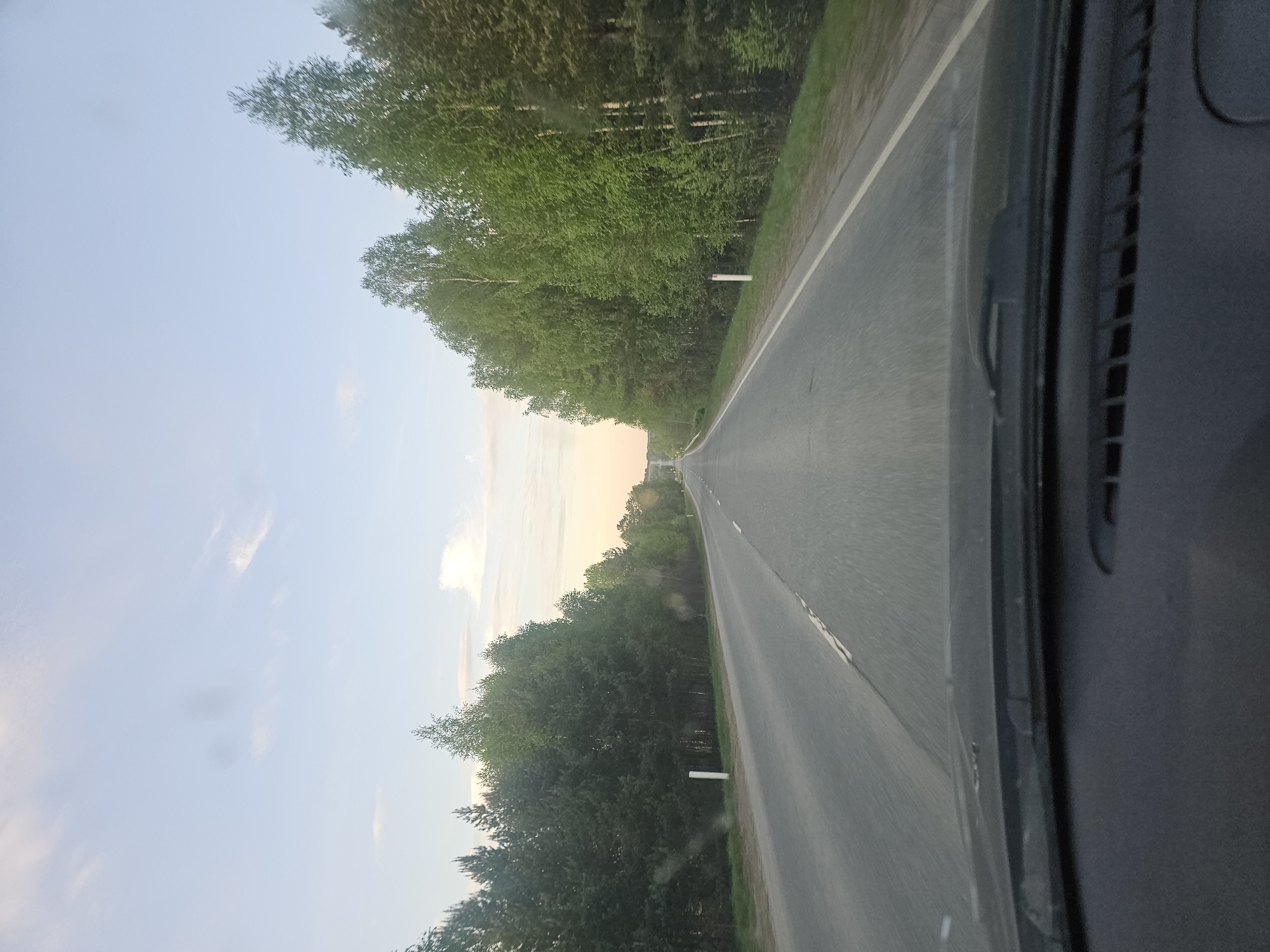 The image shows a view from inside a moving vehicle traveling on an asphalt road. The road is flanked on both sides by dense green trees. The sky is visible above the trees and appears to be light blue, potentially indicating early morning or late evening. The perspective suggests the photo was taken through the windshield of the vehicle. A portion of the vehicle's interior is visible in the right side of the frame. The location of Kirillovka, Russia, is not apparent in the image.
