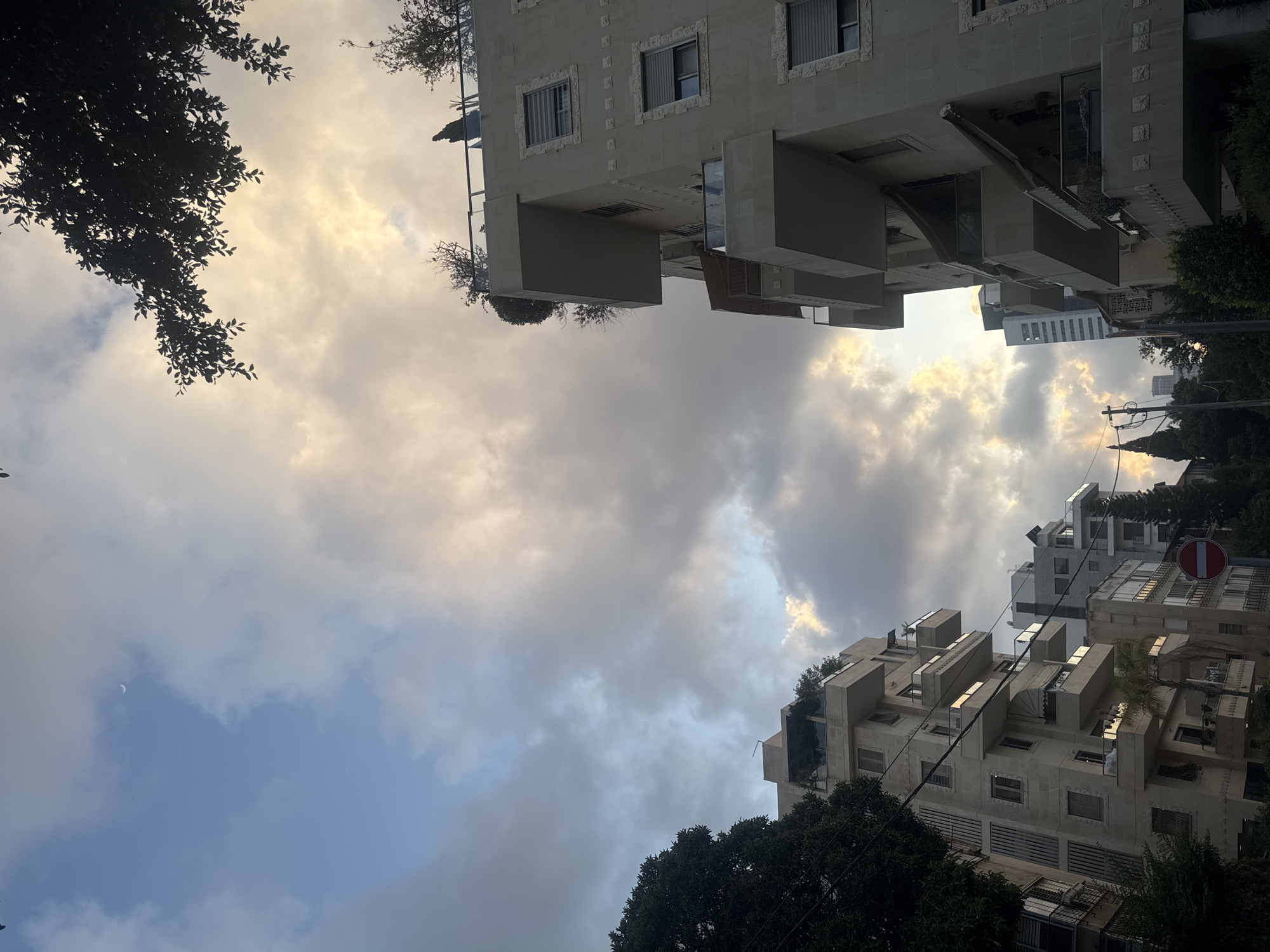 Looking upwards at an angle, the photograph captures an urban scene in Ramat Gan, Israel. The sky is filled with clouds and a sliver of a crescent moon is visible. Trees frame the view on the left and at the bottom right. Modern buildings with distinctive architectural details like protruding balconies and rooftop foliage occupy the top and bottom right quadrants of the image. A red and white traffic sign is visible on the lower right. Power lines stretch across part of the scene.