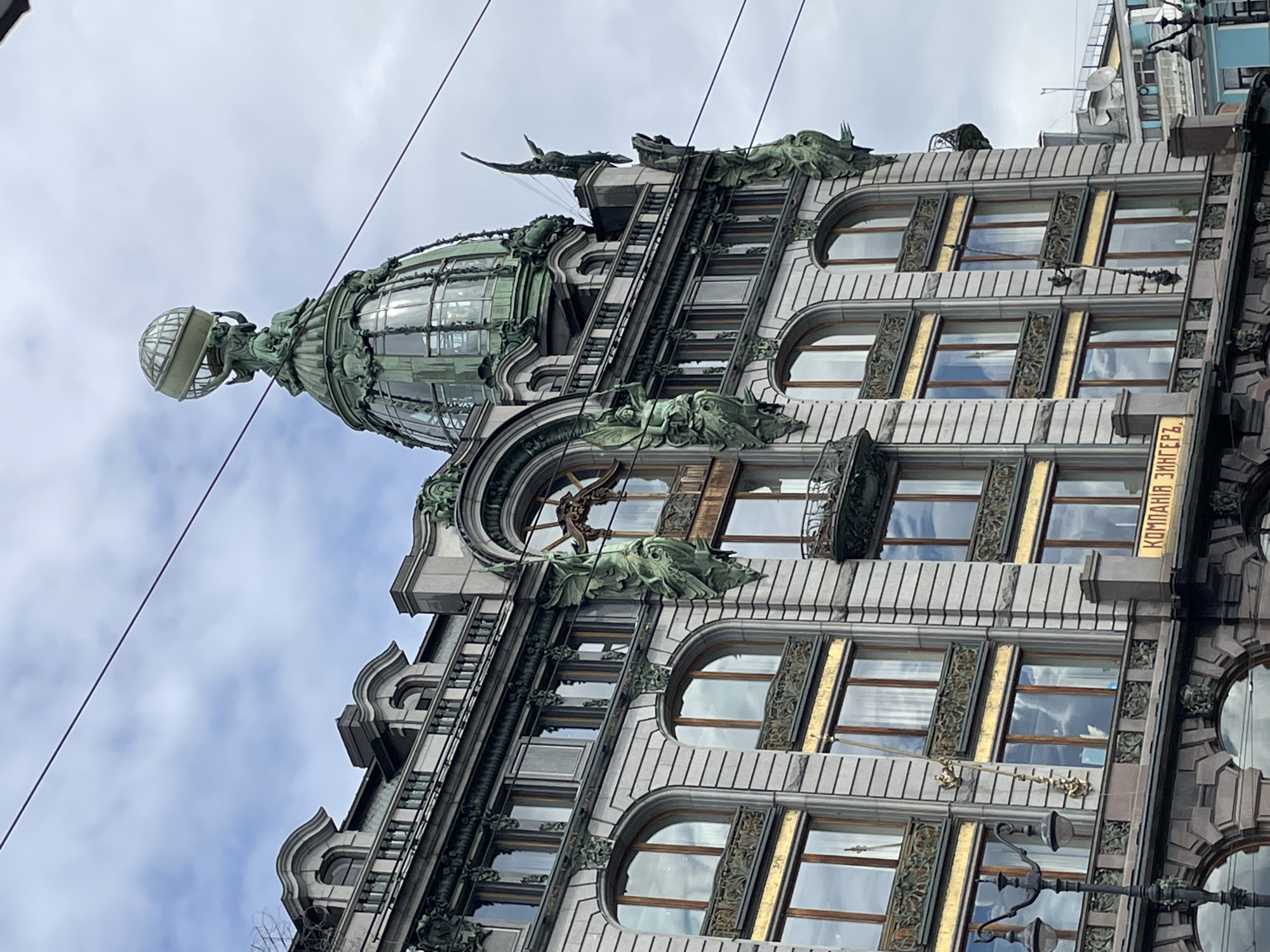 This media file showcases the Singer House, also known as the Dom Knigi (House of Books), a historic Art Nouveau building in Saint Petersburg, Russia. The image focuses on the upper floors of the building, highlighting its distinctive architectural features.
The primary subject is the building itself, characterized by its ornate facade. Notable details include a large green glass dome topped with a globe supported by an eagle sculpture. The facade is adorned with intricate sculptures of mythological figures and stylized ornaments in bronze, which have developed a green patina over time. The windows are framed with decorative golden elements, and the building displays a rich grey stone cladding.
The scene is set against an overcast sky, suggesting it is daytime. The weather appears cloudy. The image is taken from a low angle, looking upwards at the building, emphasizing its grandeur and height. There are visible power lines crossing the frame, indicating an urban setting.
The only visible text is on a yellow sign on the right side of the building, which reads "КОМПАНИЯ ЗИНГЕРЪ" (Singer Company) in Cyrillic.
There are no people or specific activities depicted in the image. The focus is solely on the architectural artistry of the Singer House.