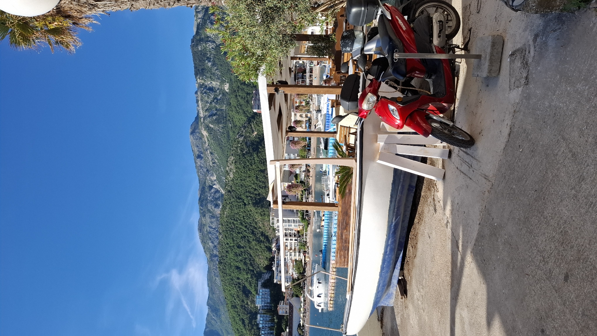 The image captures a street scene in Budva, Montenegro. A red scooter and a gray scooter are parked along the edge of a concrete road. A white boat with wooden accents is situated beside them. Behind these objects, a structure with white awnings and wooden pillars overlooks the city and the sea. Buildings are visible below, leading to the waterfront where boats are docked. A large mountain covered with trees forms a backdrop, and a clear blue sky is overhead. Palm trees are located in the upper left corner of the image.