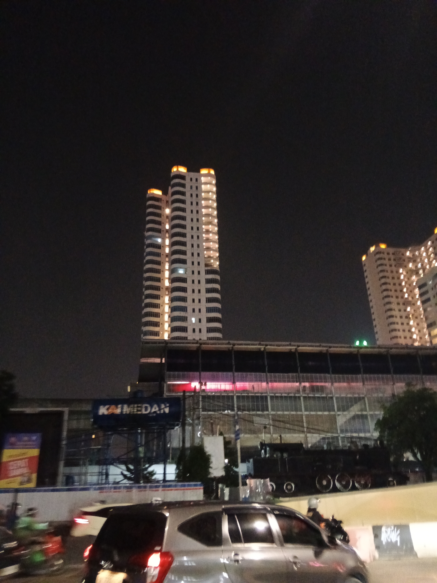 At night, a view of the City of Medan, Indonesia, shows the KAI Medan train station. The station has a modern facade with illuminated signage, and behind it, a tall, modern white building with illuminated windows stands out against the dark sky. To the right of this building, another similar building is partially visible. In front of the station, a historic black steam train is on display. The scene is busy with traffic, including cars and motorcycles, suggesting it is a public thoroughfare. The lighting suggests evening or nighttime conditions.