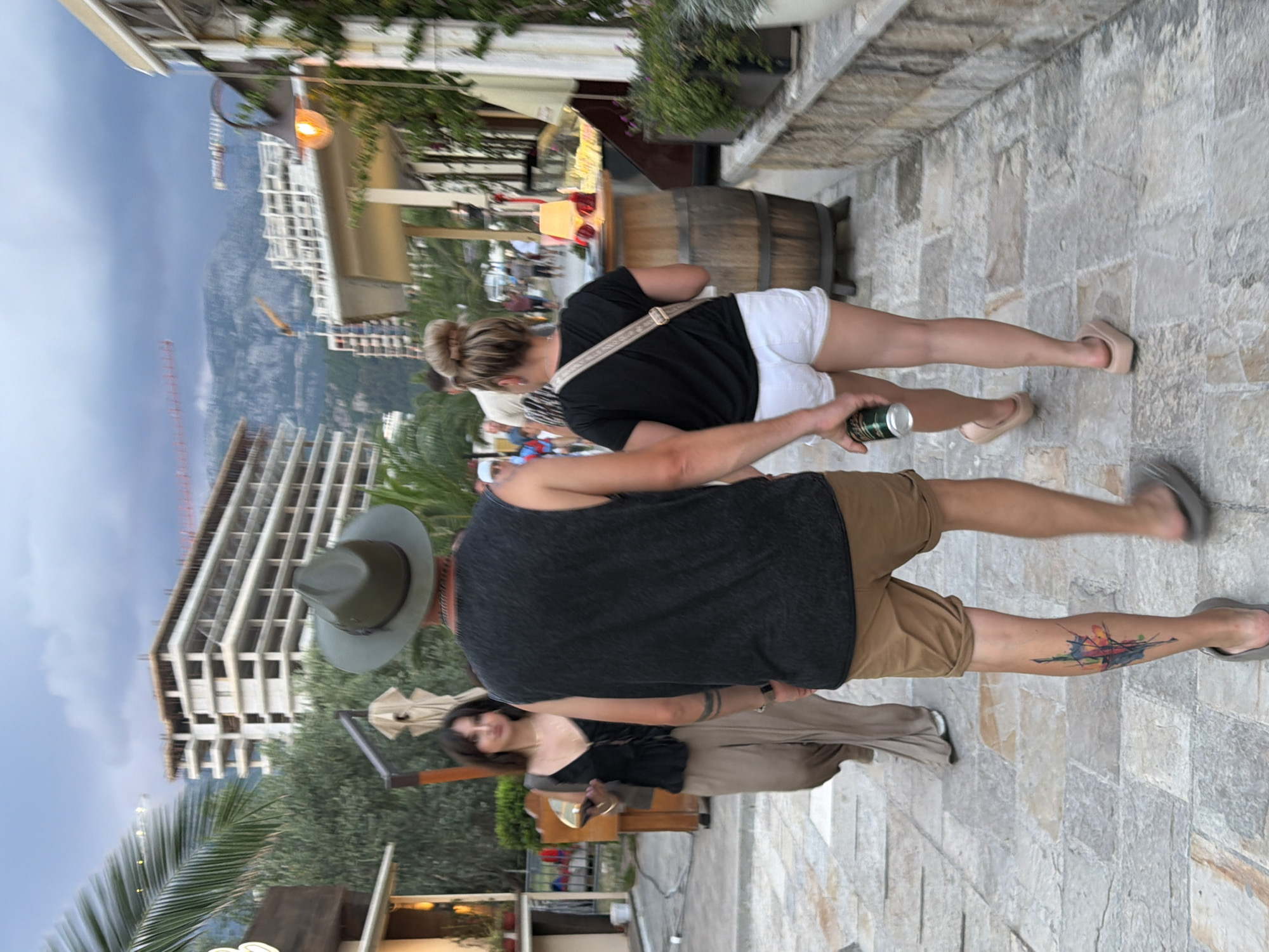 This media file shows a group of people walking on a stone-paved walkway. In the foreground, a man wearing a dark tank top, tan shorts, and a fedora hat is seen from behind. He has a colorful abstract tattoo on his left calf. To his right, a woman in white shorts and a black t-shirt is walking away from the camera. She has a crossbody bag and is holding a can. Further in the background, another woman in a black top and tan skirt is partially visible, looking towards the camera.
The scene is set outdoors with a cloudy sky and a mountainous landscape in the distance. Several buildings, some under construction, are visible on the mountainside. Lush green trees and palm fronds are also present, suggesting a warm climate. A wooden barrel and a display case with what appears to be food items are located to the left of the main subjects. The lighting suggests it might be late afternoon or early evening. The overall mood appears casual and relaxed, with people enjoying a walk. The location is identified as Budva, Montenegro. No visible text is present in the image.