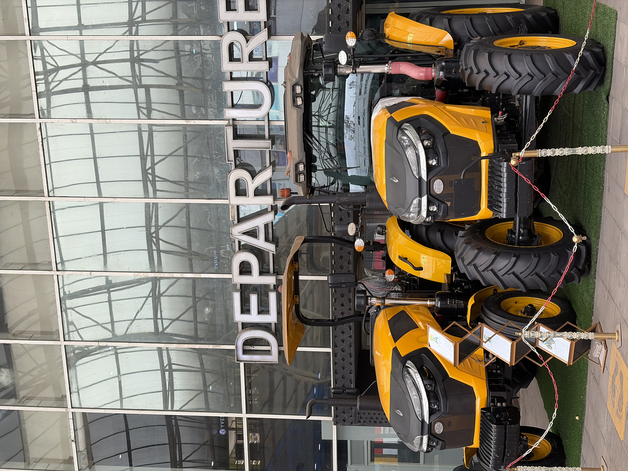 The image shows two yellow and black tractors parked in front of a building with a large "DEPARTURE" sign. The building has a modern glass facade with a visible grid structure. Each tractor is equipped with large, knobby tires and is secured by a decorative chain fence. The foreground shows artificial green grass, while the background features the reflection of the building's surroundings in the glass windows. The location appears to be a public space or an event venue, based on the display of the tractors. The city mentioned, Ikeja, Nigeria, is referenced as context, and as such may be the location where this scene exists.